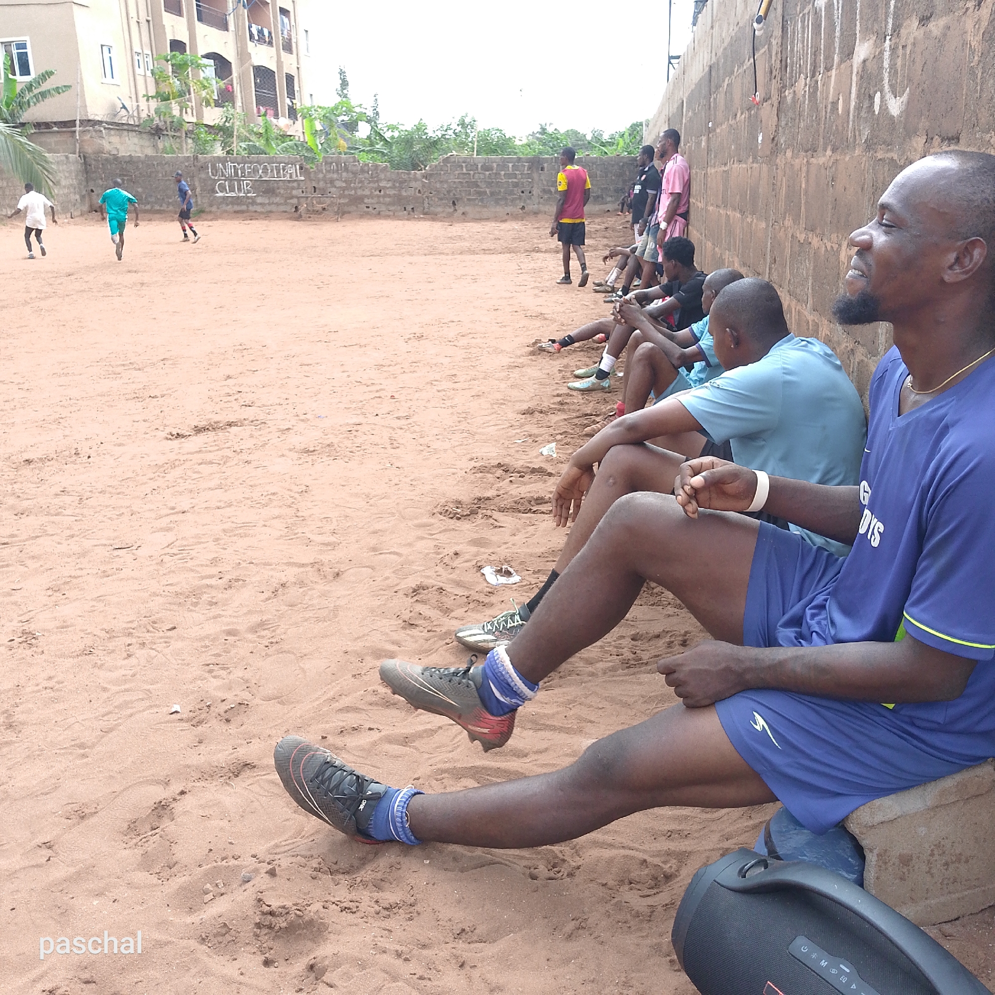 In Ukwu-Abwa, Nigeria, a dirt field serves as a soccer pitch. Several men are seated on the sidelines watching other men play soccer. A building stands behind one end of the pitch, and a block wall behind the other. A boombox sits on the ground near the seated men.