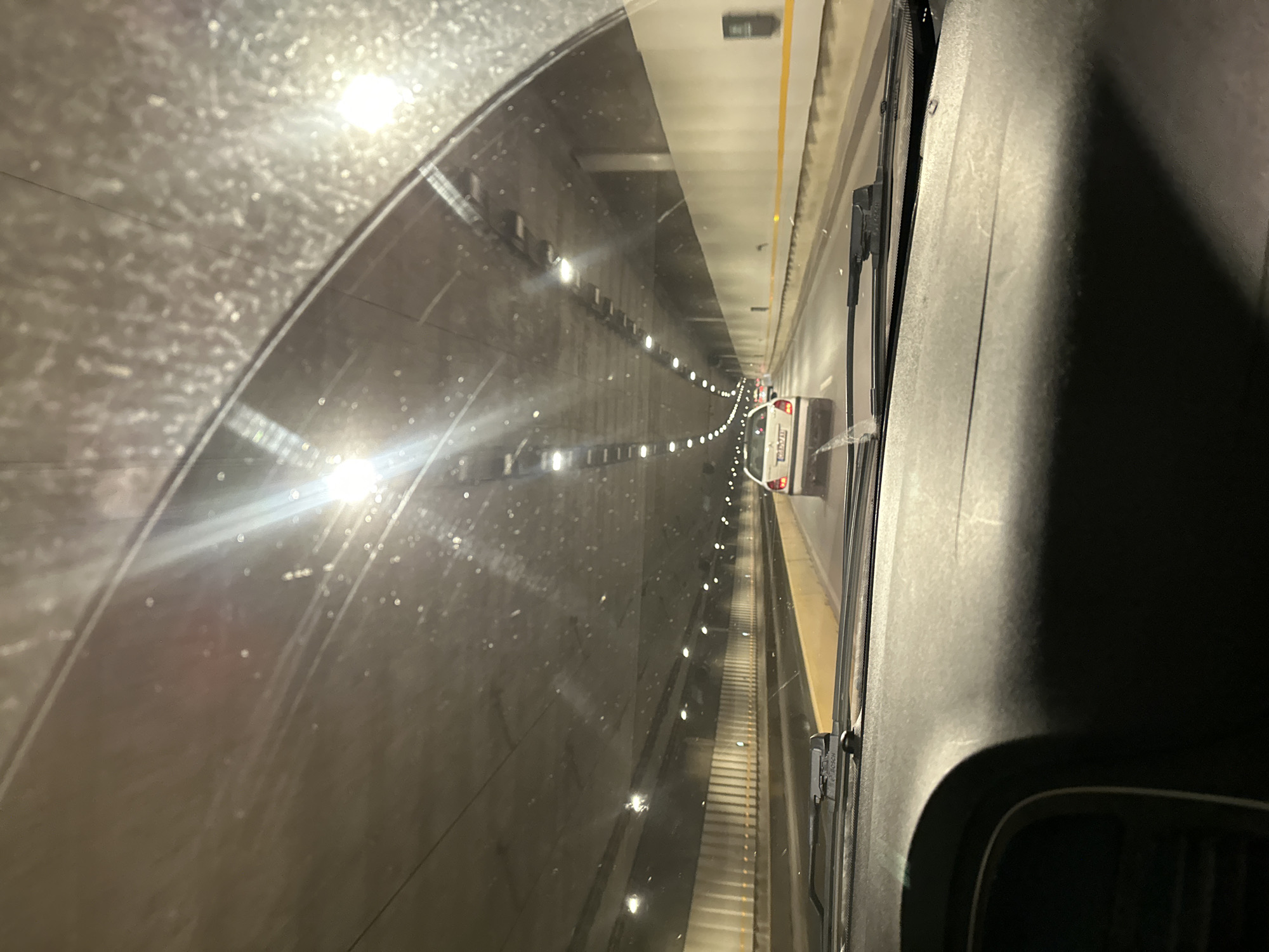 The image shows the interior of a car driving through a tunnel. The view is from the driver's perspective, looking through the windshield. The tunnel has a concrete ceiling and walls, illuminated by numerous lights. There are other cars visible in the tunnel, including a white car directly in front and a few further ahead. The dashboard of the car is visible in the foreground, and the windshield is speckled with dust or water droplets. The overall scene depicts a typical driving experience within an urban tunnel environment.