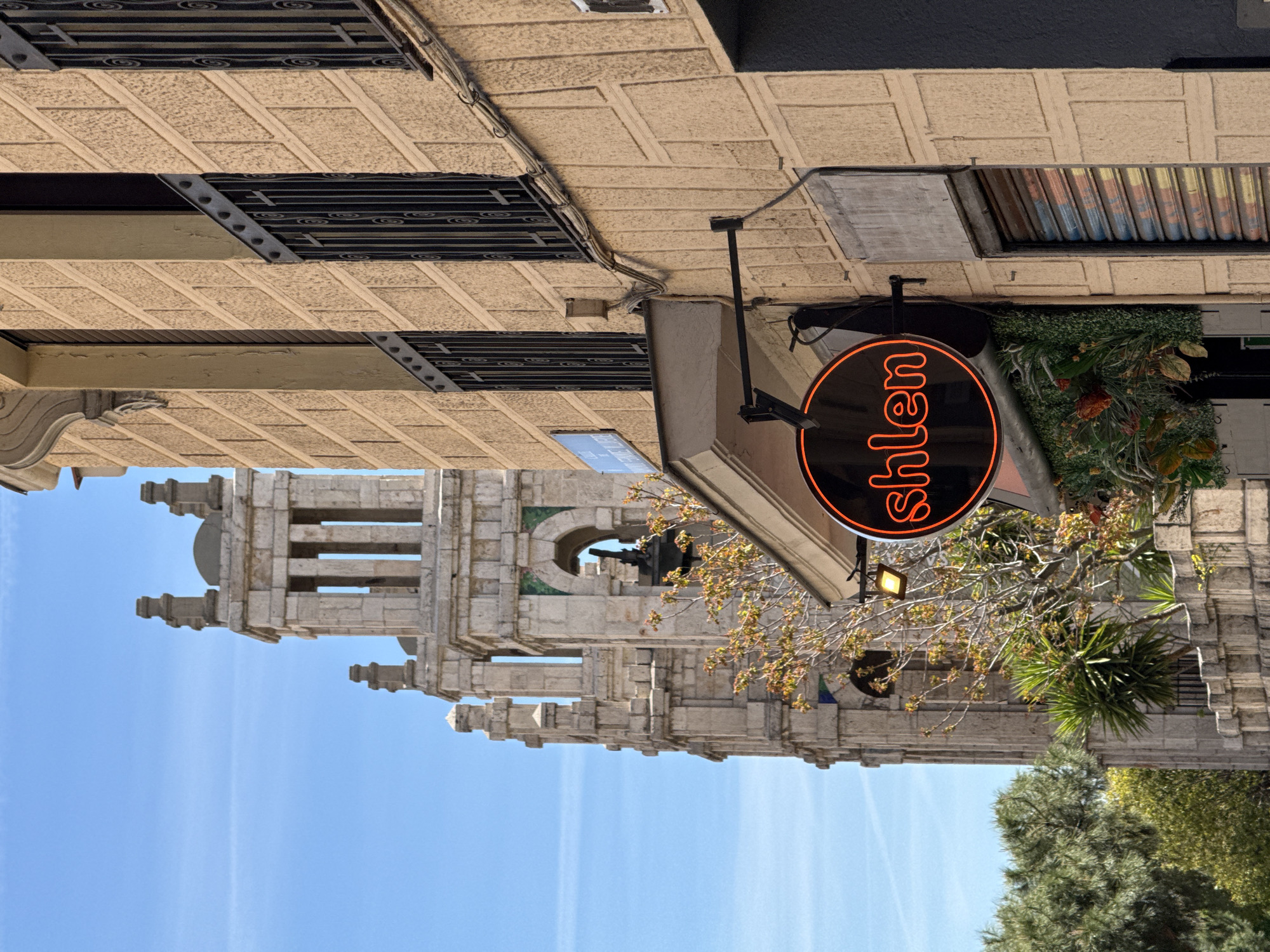 A street-level view in Madrid, Spain, shows a pale stone building with architectural details reminiscent of a church or historic structure under a bright blue sky. The foreground features a storefront with a round, black sign illuminated with orange neon lettering that reads "hlen" in a vertically flipped orientation. Above the storefront, the building facade displays beige square tiles, black grates over openings, and various conduits and fixtures. To the right of the sign, lush greenery cascades down the building. Trees and foliage are also visible in the lower right corner of the frame.