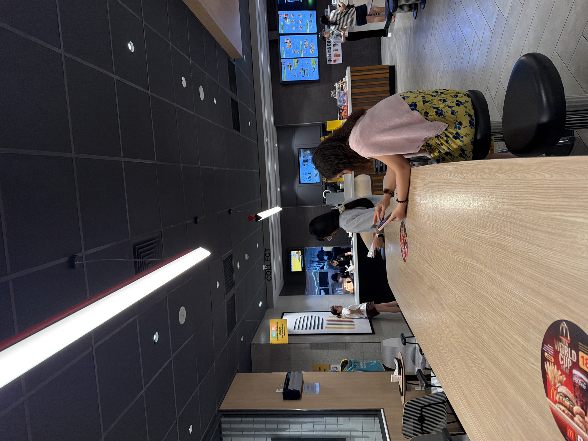 This image depicts the interior of a McDonald's restaurant in Đà Nẵng, Vietnam. The scene is captured from a slightly elevated perspective, looking down and across a long wooden counter. Several people are visible, primarily staff members behind the counter and customers on the other side.
In the foreground, a person wearing a pink top and a yellow floral skirt is leaning over the counter, looking at their phone. To their left, another person, possibly a customer, is seated. Behind the counter, several employees are attending to orders. One is holding a phone, and others are engaged in food preparation or serving. In the background, digital menu boards display food items and promotions. One of the boards has Vietnamese text, including "Quét mã QR" which means "Scan QR Code." Another sign reads "Khu vực dành cho đại đơn hàng," translating to "Area for large orders."
The setting is a modern fast-food restaurant, with clean lines, a tiled ceiling, and bright fluorescent lighting. The overall atmosphere appears to be a typical busy lunchtime or evening at a popular eatery. The presence of "WORLD CUP MEAL" branding suggests the photo might have been taken during a World Cup event. The image is oriented vertically, with the top of the scene appearing on the left.