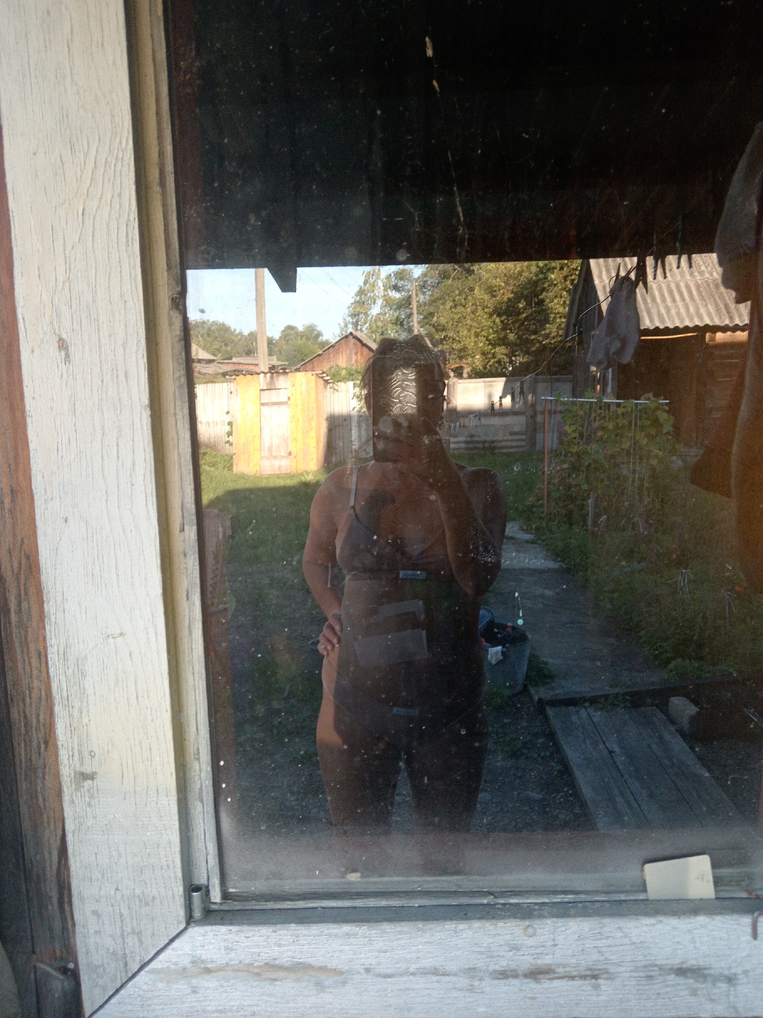 A woman's reflection is visible in a dirty window pane, likely from a rural dwelling. The woman is wearing a dark swimsuit or lingerie and appears to be taking a selfie. Her face is obscured by the camera. Behind her, through the window, a backyard scene is depicted with trees, a wooden fence, and a shed with a metal roof. The overall impression is of a casual, outdoor setting in a rural area. The context of Novopokrovka, Russia is not visually evident from the image itself, but could be inferred from external information.