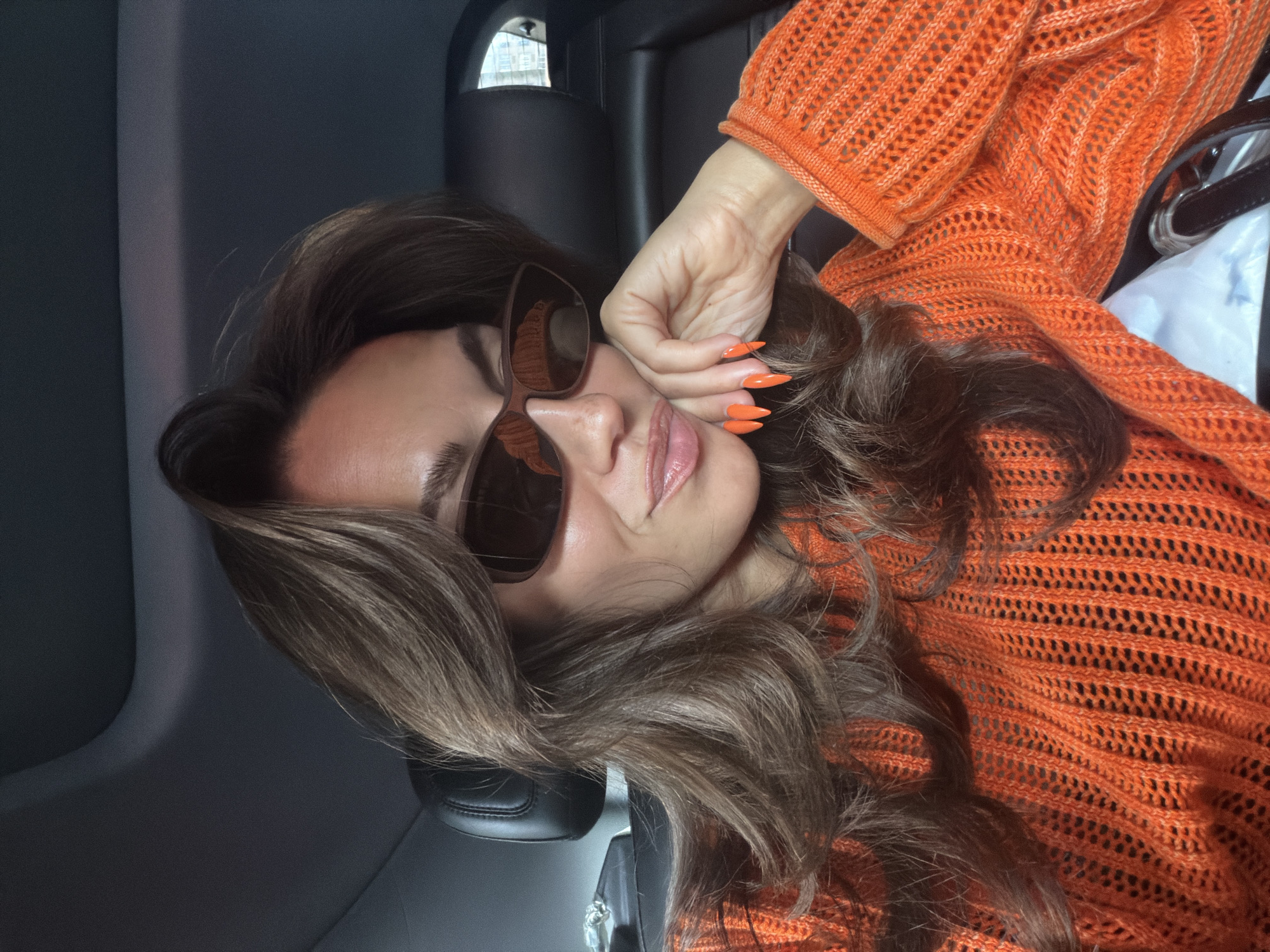 An eye-level close-up shot captures a person, likely female, posing inside a vehicle. The individual's head is tilted to the left, with long, wavy brown hair framing her face and cascading over her shoulders and an orange knitted top. She is wearing large, dark brown sunglasses that obscure her eyes, and her lips are slightly parted, revealing a hint of a smile. Her right hand, adorned with long, sharply pointed bright orange fingernails, is gently placed near her temple, partially covering her face.
The orange knitted top features an open-knit or crocheted pattern, providing a textured contrast to her smooth skin and hair. The background consists of the dark gray or black interior of what appears to be a car, with a headrest visible behind her. A reflection of a window or outer environment can be seen in the sunglasses. To the far right, a dark strap from a bag or purse is partially visible, along with some lighter material that could be a plastic bag.
The lighting suggests it is daytime. No specific location cues are visible within the image to confirm "New York, United States of America," beyond the generic car interior. The overall mood conveyed is relaxed and stylish. There is no visible text in the image.