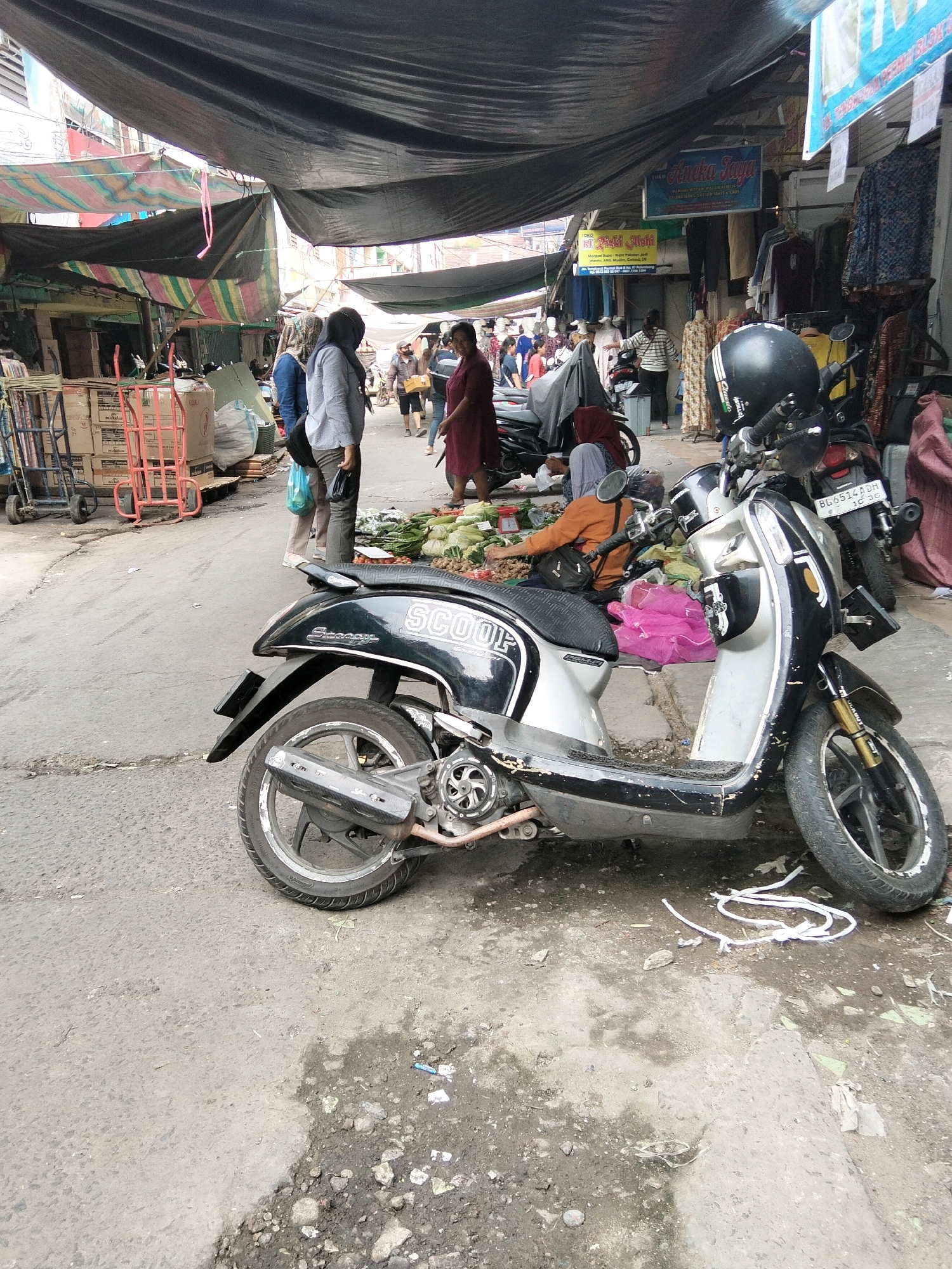 The image depicts a street market scene in Palembang, Indonesia. A black and silver scooter, with the word "SCOOP" visible on its side, is parked in the foreground, angled towards the right. The scooter is adorned with a black helmet and has a pink bundle on its seat. Behind the scooter, several people are visible. On the left, two individuals, one wearing a light blue shirt and the other a gray top, are standing near a pile of boxes and red carts. Further back, a woman in a maroon dress is seen walking. A person wearing an orange top and a headscarf is seated near a display of vegetables, including corn and leafy greens. The market stalls, covered with dark fabric and colorful tarps, are lined up along the street. The ground is paved with asphalt and appears somewhat littered. The overall impression is of a bustling, everyday market environment.