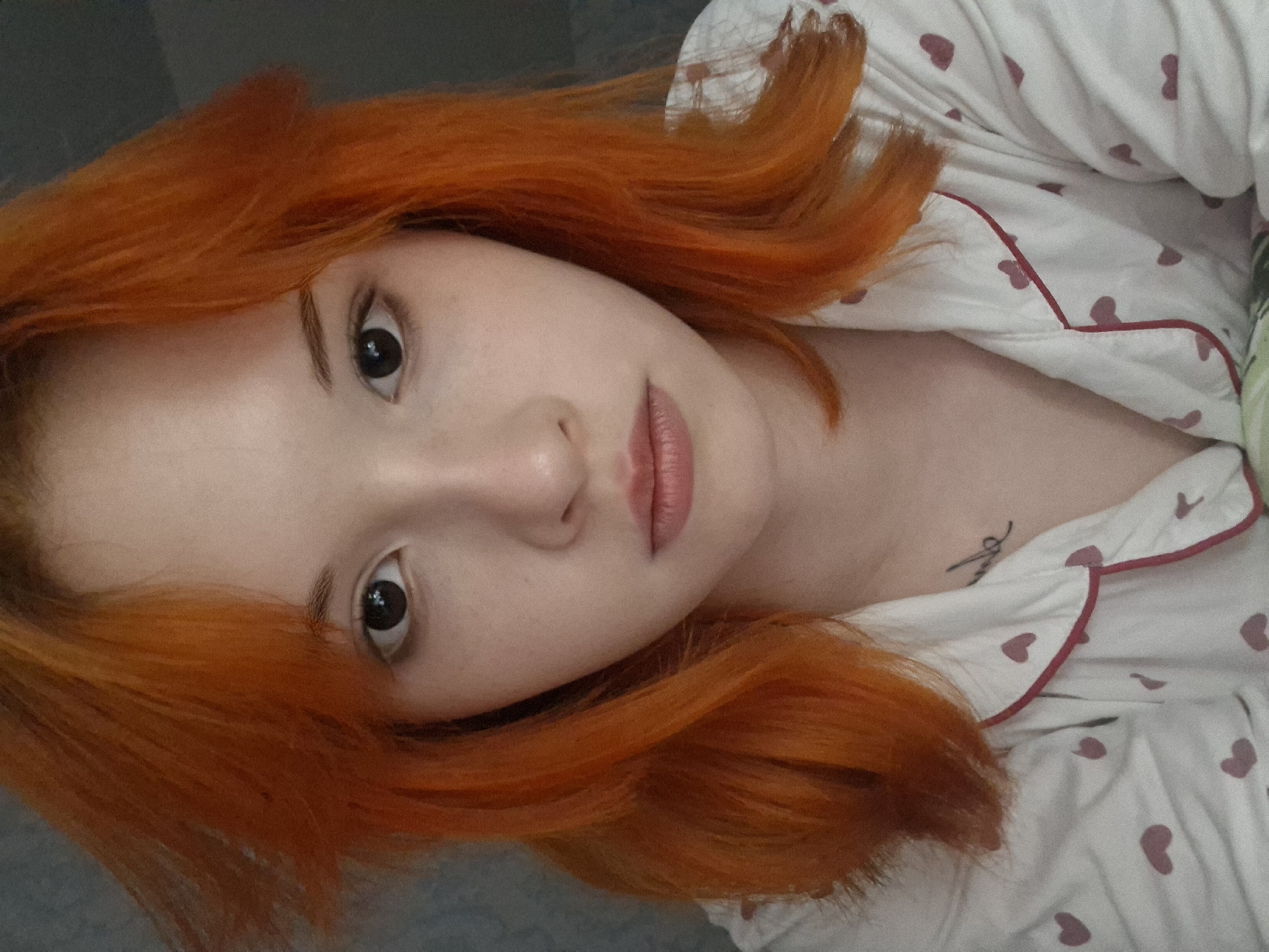 This media file features a close-up portrait of a young person, likely female, with striking, vibrant orange hair and a fair complexion. She is looking directly into the camera with large, dark eyes and a neutral, calm expression. Her head is tilted to the side, suggesting she is reclining or lying down.
She is wearing white pajamas adorned with small, repetitive pink heart patterns and thin red piping along the edges of the fabric. A small, stylized tattoo is visible on her upper chest or collarbone area; its specific text or design is not clearly discernible. The background is indistinct and appears as a muted, dark grey tone, suggesting an indoor setting, possibly a bedroom. The lighting is soft and even, highlighting her features and the texture of her hair.
The image conveys a sense of casual intimacy, typical of a selfie or informal portrait. The location context provided is Chaikovsky, Russia, though no specific visual cues within the image itself confirm this.