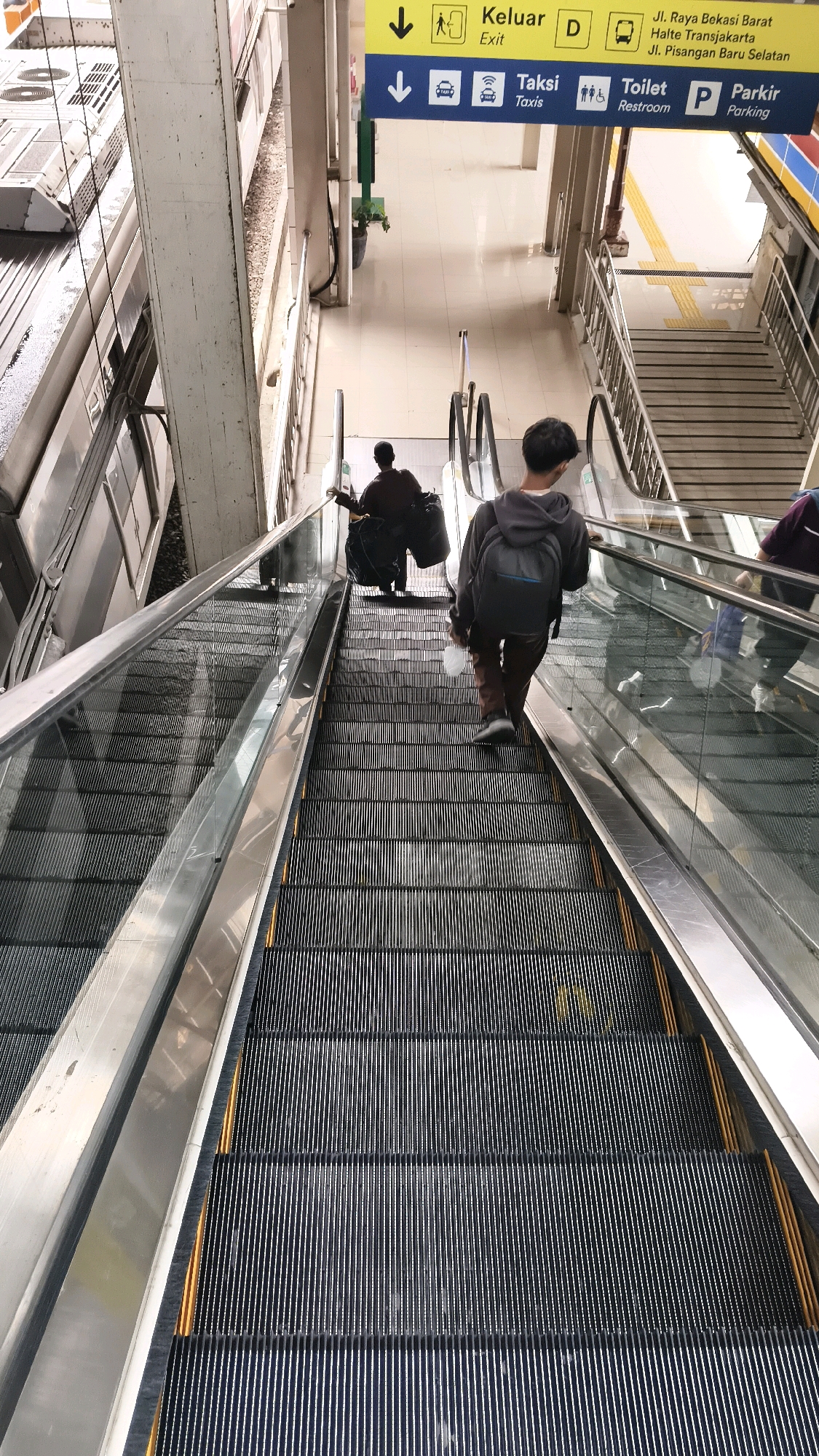 The image captures an escalator descending at a public transportation hub, likely a train station, in East Jakarta, Indonesia. The escalator's steps are metallic and black, bordered by a bright yellow safety strip and a polished metal handrail. Above the escalator, a blue and yellow directional sign is visible. The sign contains text in Indonesian and English, indicating "Exit," "Taksi" (Taxis), "Toilet Restroom," and "Parkir Parking," as well as street names "Jl. Raya Bekasi Barat" and "Jl. Pisangan Baru Selatan."
Two individuals are descending the escalator. In the foreground, a person wearing a grey hoodie and dark pants, carrying a grey backpack, is walking down. They are holding a white plastic bottle in their right hand. Further up the escalator, another person, facing away from the camera, is also descending. They are wearing dark clothing and carrying two large, dark bags.
To the left of the escalator, a train carriage is partially visible, suggesting the location is adjacent to a railway line. The overall environment appears to be indoors, with a tiled floor and visible support structures. The lighting suggests it is daytime.