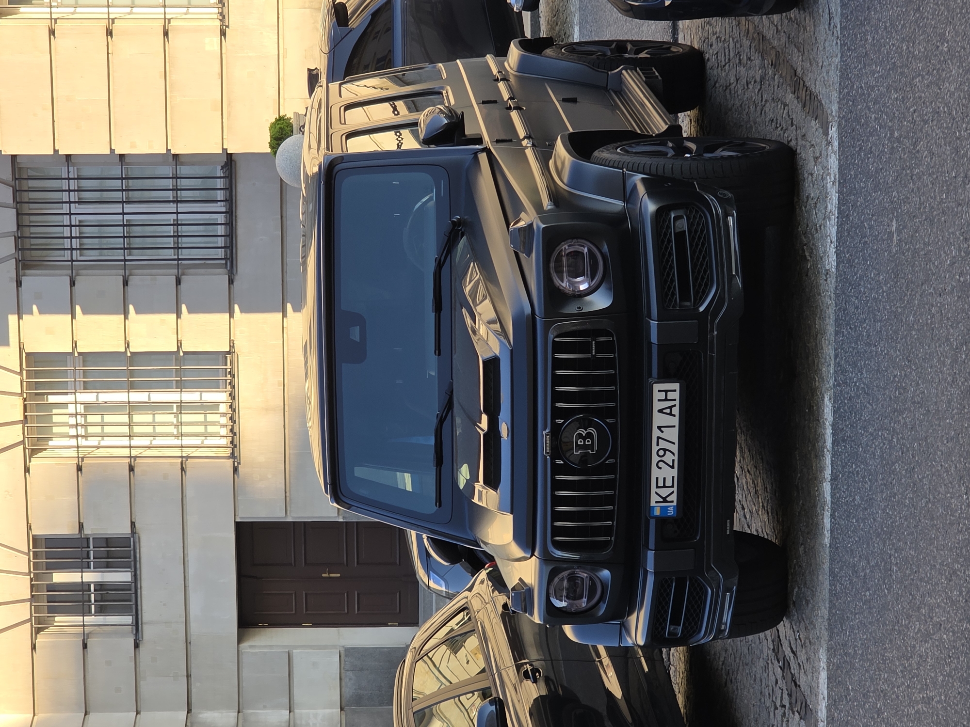 An exterior, eye-level, full shot captures a matte black Brabus G-Wagen parked on a cobblestone street in Warsaw, Poland. The vehicle is angled slightly towards the camera, with its front grille and headlights prominently displayed. The Brabus emblem, a stylized 'B', is visible on the grille. The car's license plate, bearing the Ukrainian characters "UA KE 2971 AH," is clear. A portion of another dark-colored vehicle is visible to the left of the Brabus. In the background, the facade of a building features large windows with metal bars and a dark brown double door. The lighting suggests it is late afternoon, with long shadows cast on the building and street, indicative of a sunny day. The scene is static, with no people or active interactions depicted.