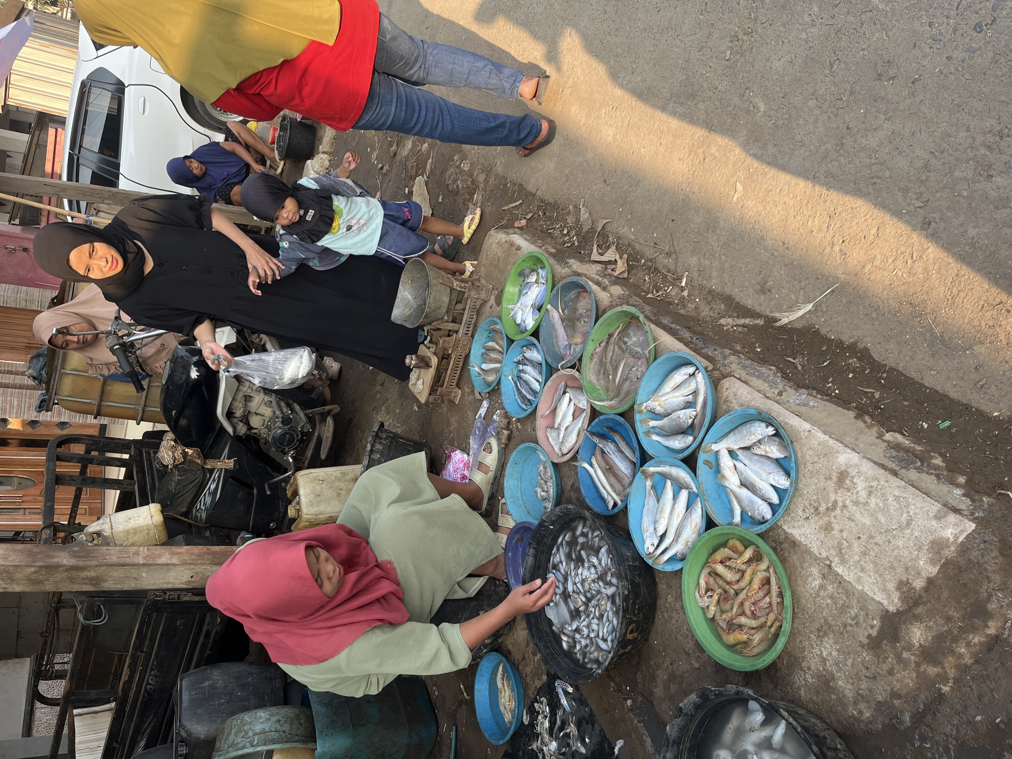 In Kedungmalang, Indonesia, a woman wearing a pink hijab and a green outfit is seated in front of several containers of fish and shrimp at what appears to be an outdoor market. To her right, a woman in a black hijab and dress stands, holding a plastic bag and a young child wearing a blue shirt and shorts. Other individuals, including a woman in a blue hijab and a woman in a yellow hijab, are also present, along with a white car and a motorcycle. The ground is dirt and concrete, with various plastic containers and scales scattered around. The scene is illuminated by sunlight casting long shadows.
