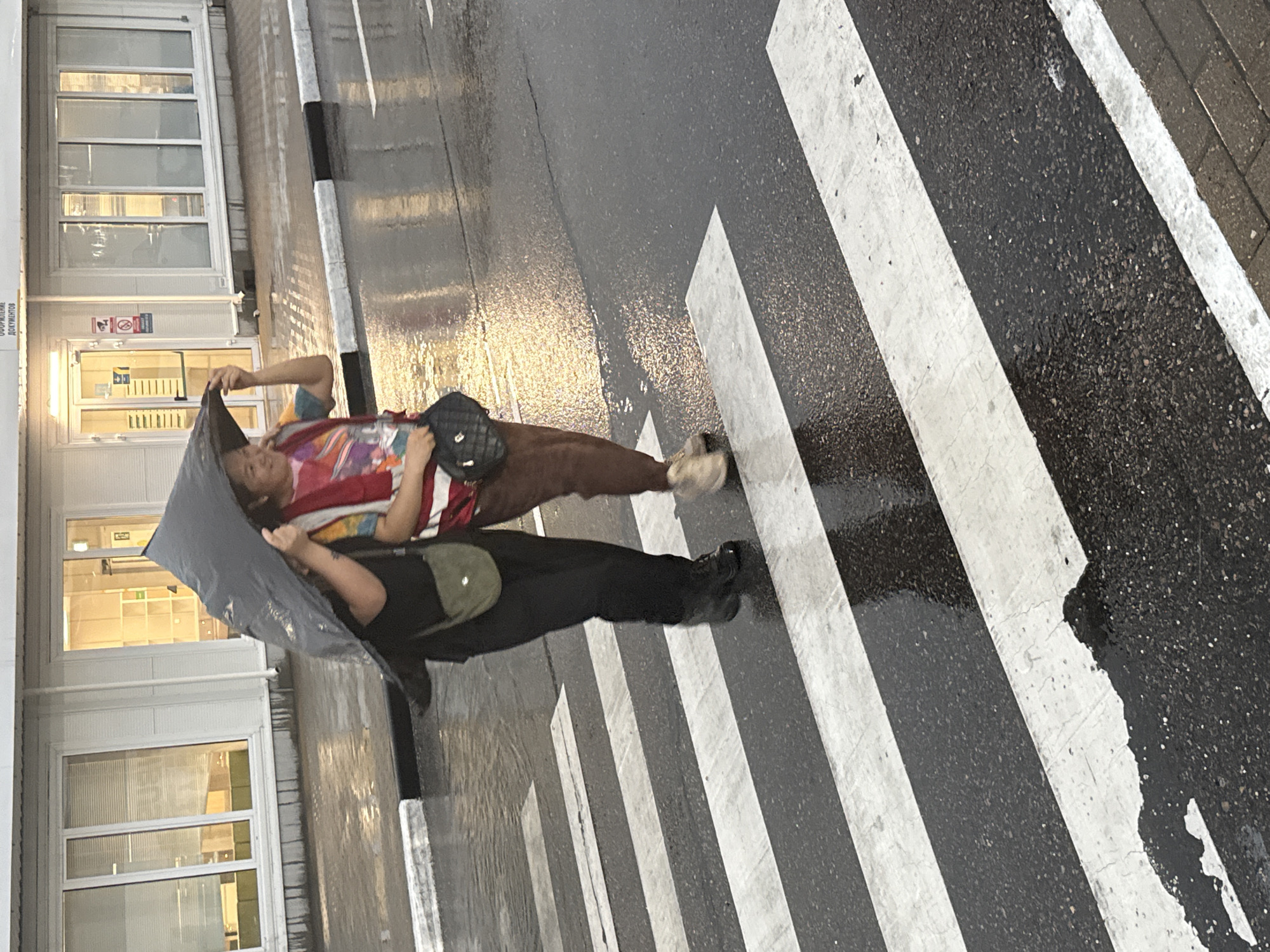 Two individuals are crossing a wet, striped crosswalk in Lupanovo, Russia. The person on the left is wearing black pants, a black top, and a light green crossbody bag, holding a large, dark umbrella over both individuals. The person on the right is wearing brown pants and a colorful, possibly tie-dye, shirt and red vest, and is carrying a black purse. The ground is wet and reflective, indicating recent rainfall. In the background, there is a building with large windows and a doorway. The overall scene depicts a rainy day in a town setting.