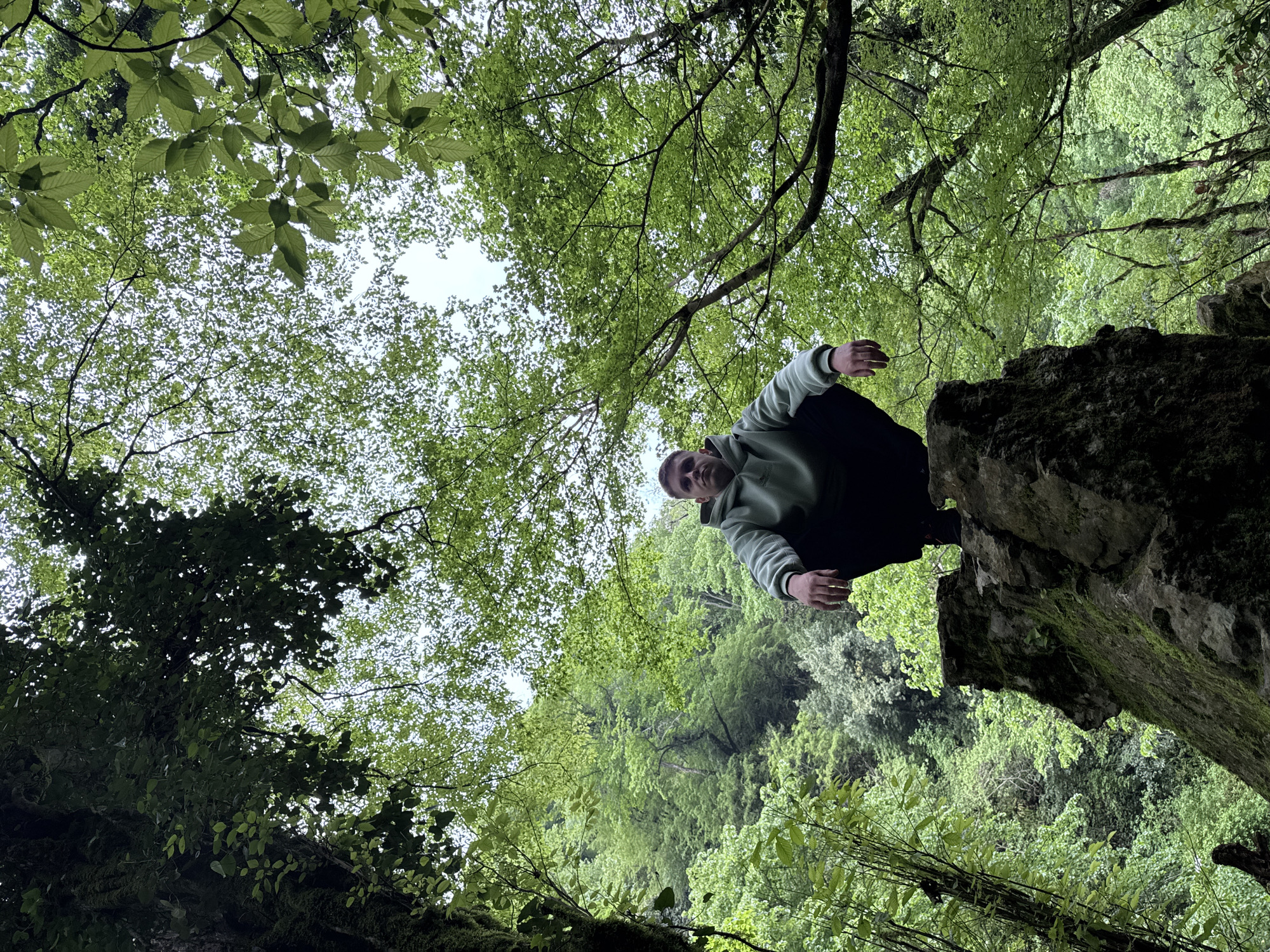 A low-angle shot depicts a person perched atop a moss-covered rock formation, with a dense forest canopy overhead. The person, wearing a light-colored hooded sweatshirt, dark pants, and a dark beanie, squats on the rock, looking directly down at the camera. The forest canopy fills most of the background, composed of green leaves. The location is inferred to be the city of Tsabal in Abkhazia.