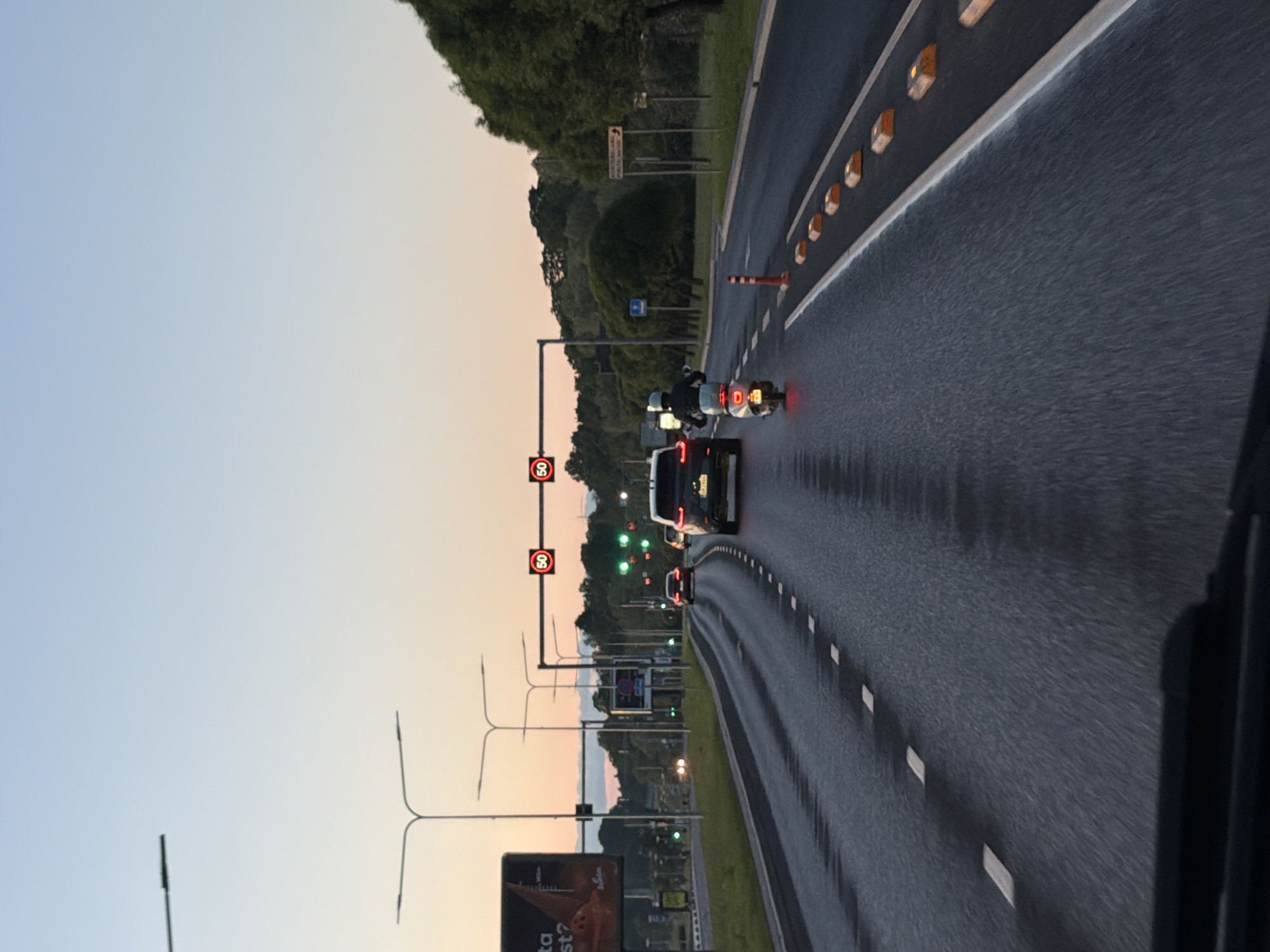 A street-level, eye-level, vertical shot shows a view from inside a vehicle on a multi-lane road. The road is wet and has dashed white lines marking the lanes. In the foreground, the top edge of a car's dashboard is visible. The scene depicts a slight dusk or dawn sky with hues of light blue and pale orange.
Several vehicles are on the road. A black SUV is in the right lane, and a scooter is in the left lane, with a rider wearing a helmet. Further ahead, another black SUV is visible. Traffic lights with red "50" signs are illuminated above the lanes. Streetlights and trees line the sides of the road. A billboard advertisement is present on the left side of the road.
The location is likely an urban or suburban area, potentially a highway or major road. Given the visual cues such as the road infrastructure, traffic lights, and vegetation, this could be a scene from Tallinn, Estonia.
