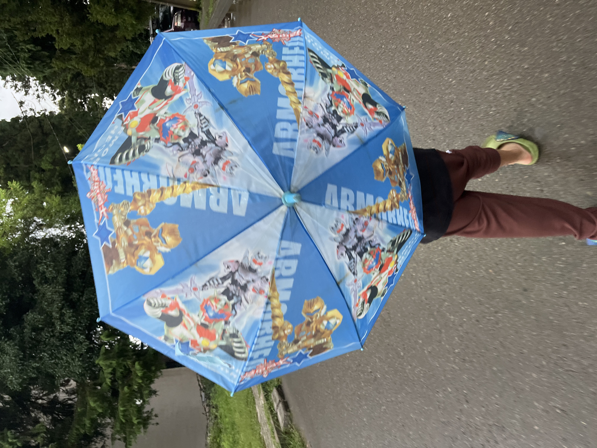 A person walks on a wet asphalt road, holding a closed blue umbrella featuring a colorful "Armor Hero" design with robots and stars. The person is wearing dark pants and light green open-toed sandals. The surroundings show a grassy area on the left and trees on the right, with a hint of a building and a parked car in the background. The scene appears to be a street in Ureki, Georgia, given the context of the request. The lighting suggests an overcast or rainy day.