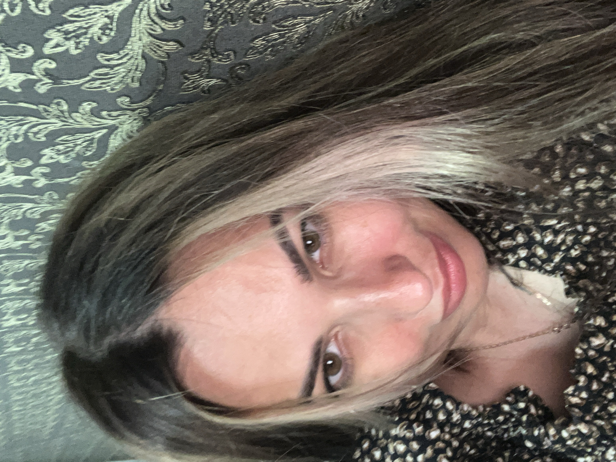 A woman is captured in a close-up selfie, her face filling most of the frame. She has brown eyes and appears to be wearing makeup, with defined eyebrows and pink lipstick. Her hair is long and wavy, with dark brown roots and lighter blonde streaks running through it. She is wearing a dark-colored top with a patterned design. The background consists of a textured wallpaper with a damask-like pattern in shades of grey and gold. The lighting is soft, illuminating her face and highlighting her features. The overall impression is that of a casual, personal photograph.