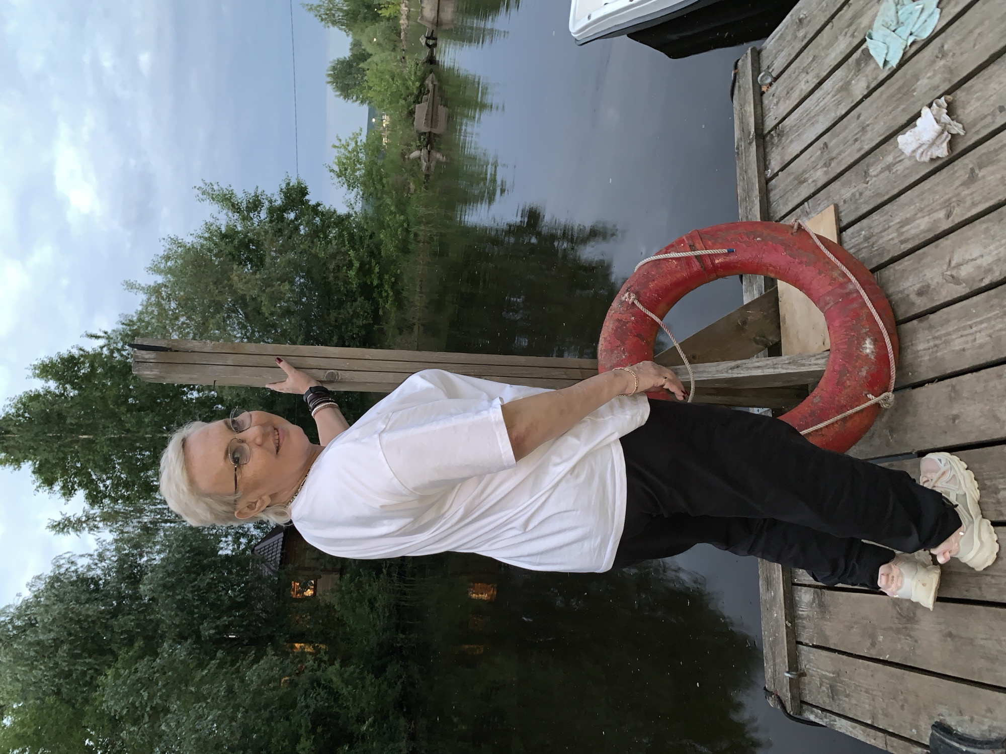 A woman with short blonde hair and glasses, wearing a white t-shirt and black pants, stands on a wooden dock. She is holding onto a wooden post. A red life preserver is on the dock next to her. A body of water is behind her, with trees on the opposite bank. A small house with a light on is visible among the trees. A white boat is partially visible on the right side of the dock. The sky is cloudy. The scene appears to be located near the Kama River in Chaikovsky, Russia, a city known for its hydroelectric power station and proximity to the river.