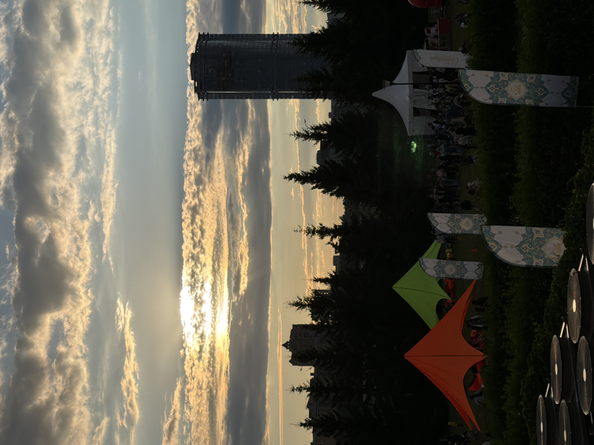 The image depicts an outdoor event at dusk in the city of Unknown, Unknown. The sky is filled with dramatic clouds illuminated by the setting sun. In the background, a tall, modern skyscraper stands silhouetted against the sky, accompanied by other buildings and a dense line of dark green evergreen trees. In the foreground, there are several brightly colored tents, including an orange and a lime green one, set up on a grassy area. A larger white tent is also visible, with a crowd of people gathered around it. Decorative flags with intricate patterns are placed throughout the scene. The overall atmosphere is that of a festival or gathering taking place as daylight fades.