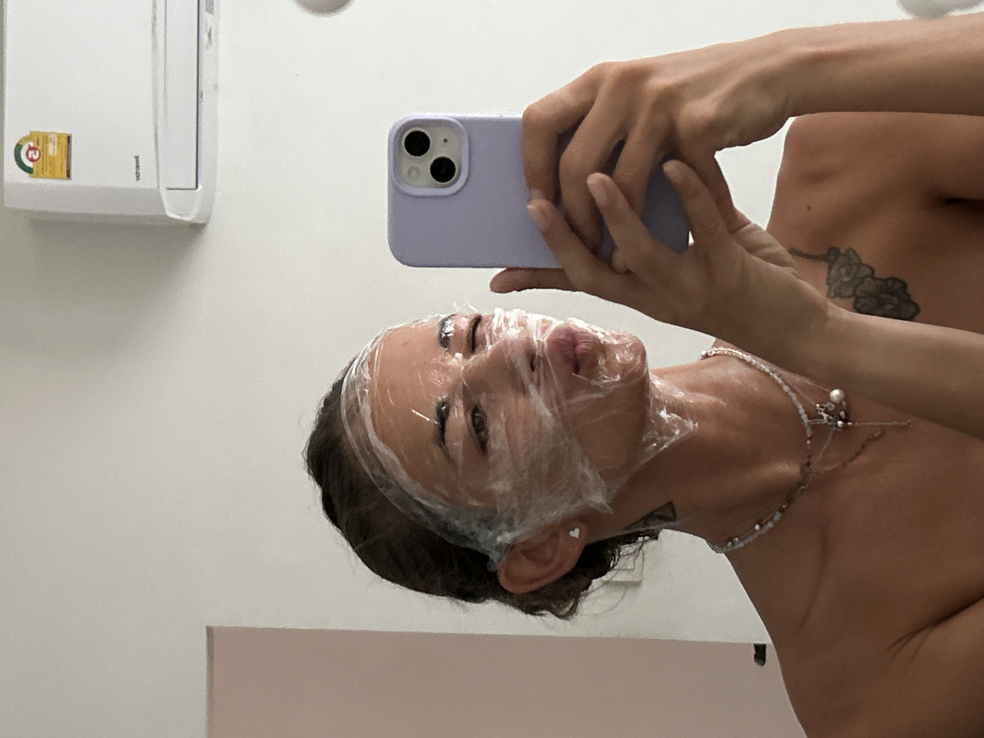 A young woman, with her hair pulled back, is taking a selfie in a room. She has plastic wrap covering her face, obscuring much of it, but her lips are puckered. She is wearing a light purple phone case. A tattoo is visible on her shoulder. In the background, a white air conditioning unit is mounted on a light-colored wall. A pink surface is partially visible on the left side of the frame. The image appears to have been taken in Kathu, Thailand.