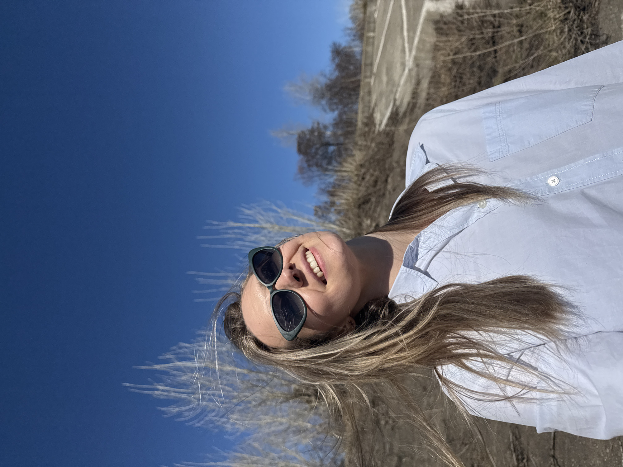 A fair-skinned woman with long, blonde hair smiles while wearing dark sunglasses and a light blue button-down shirt. She is outdoors, seemingly on a partially paved area near some bare trees. The sky is a clear, deep blue. The photograph appears to be taken in the city of Chaikovsky, Russia. The specifics of the precise location within Chaikovsky are not clear in the photo.