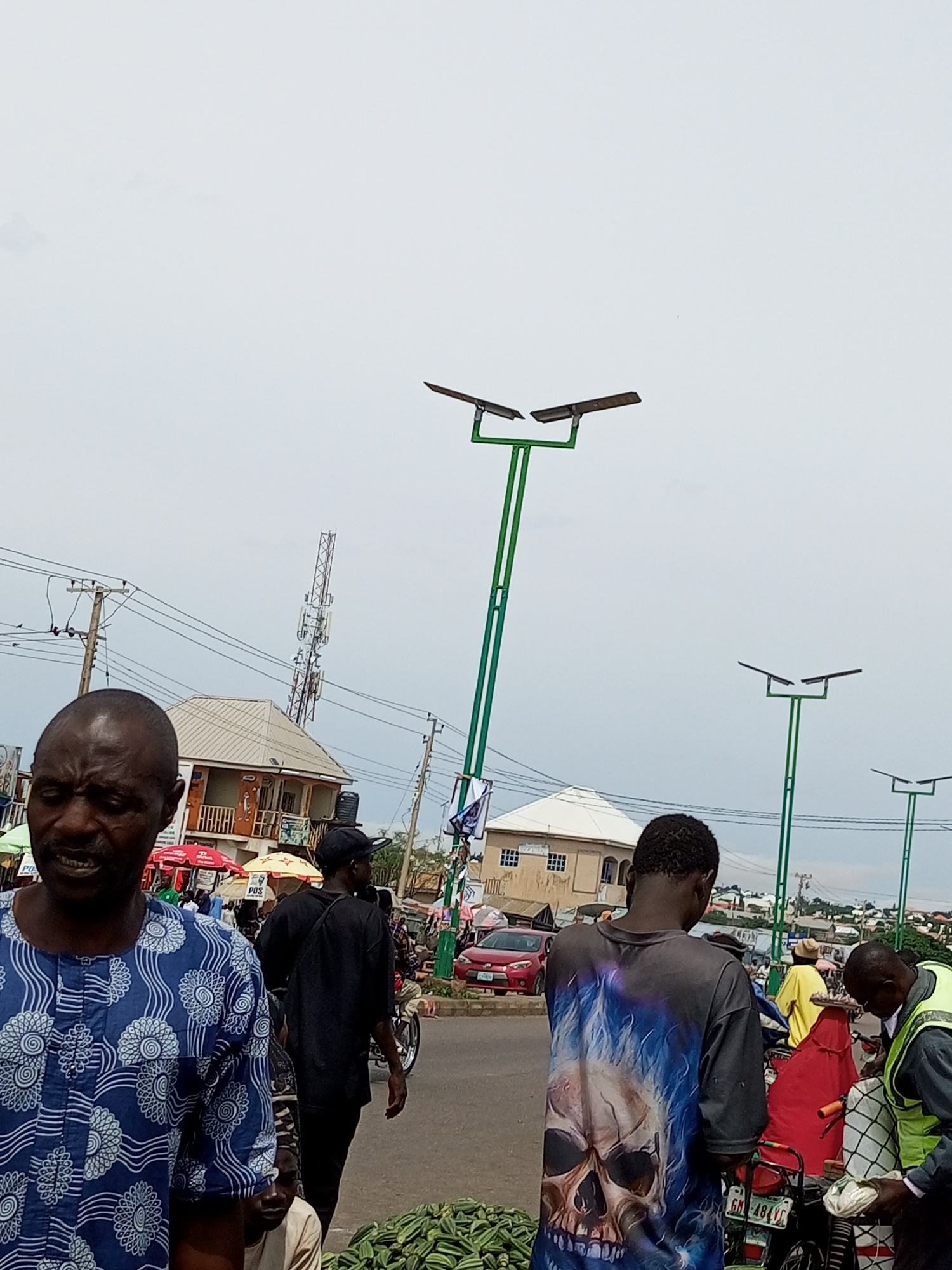 This is a full shot of a busy street scene in Tempure, Nigeria, under an overcast sky. In the foreground, a man wearing a blue and white patterned traditional outfit is looking to his left with his mouth slightly open, appearing to speak. Behind him, a person in a black t-shirt with a skull and flames graphic is facing away from the camera, and another person in a black t-shirt and cap is walking away. Several solar-powered streetlights with green poles stand tall throughout the image. In the background, there are buildings, a cellular tower, power lines, and various vehicles, including cars and a motorcycle. Market stalls with umbrellas and produce, such as okra, are visible on the side of the road. People are seen walking and interacting in the background, indicating a bustling market atmosphere.
