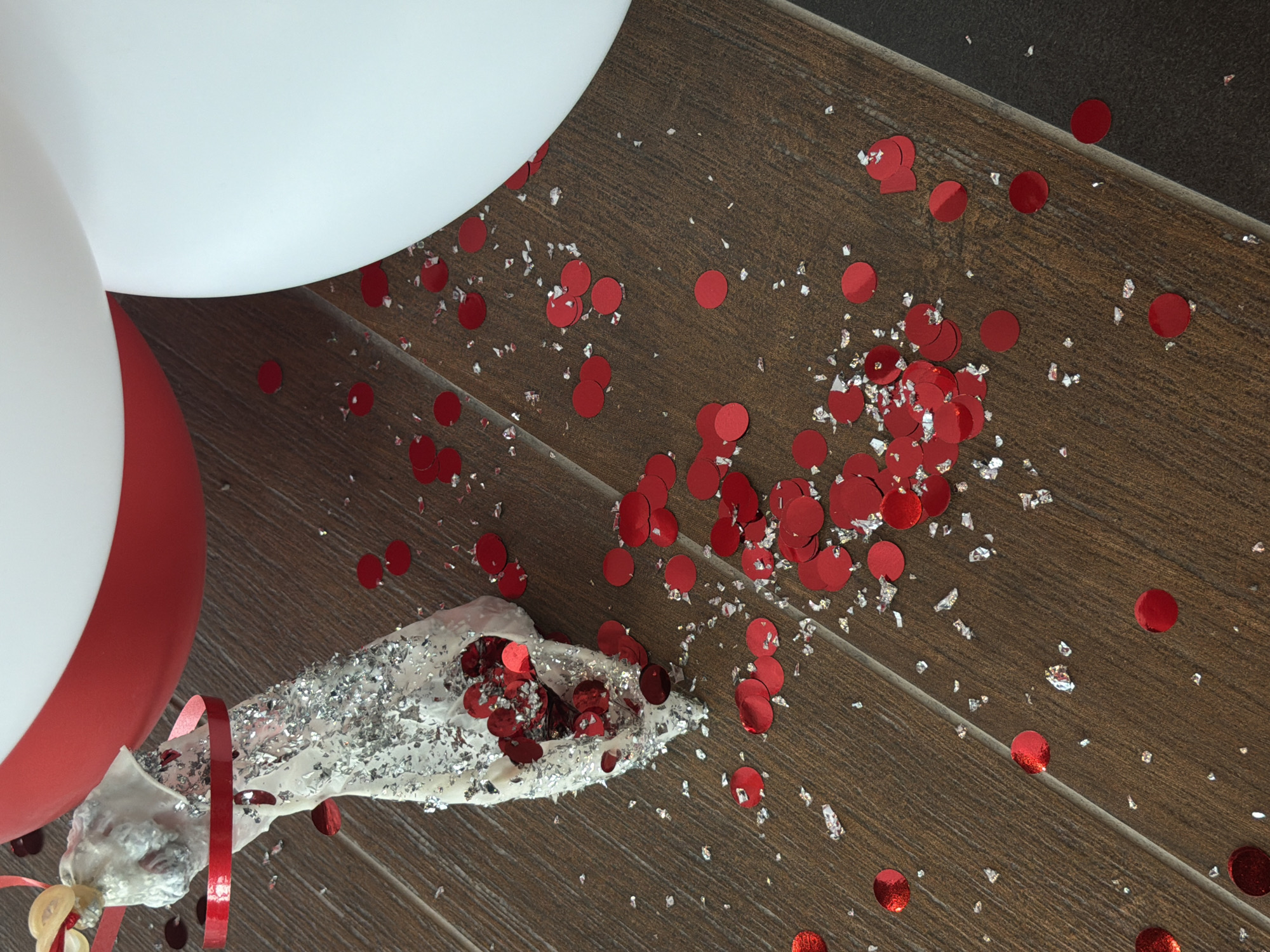 The image shows a top-down view of a scene on a wooden floor. Red confetti and silver glitter are scattered across the floor. A deflated balloon in shades of red and white rests on the floor, along with a flattened, silver-covered object, possibly a party favor or noisemaker. The image provides no information about the location in Izhevsk, Russia, people, actions, or interactions. The environment consists of the floor and scattered celebratory debris.