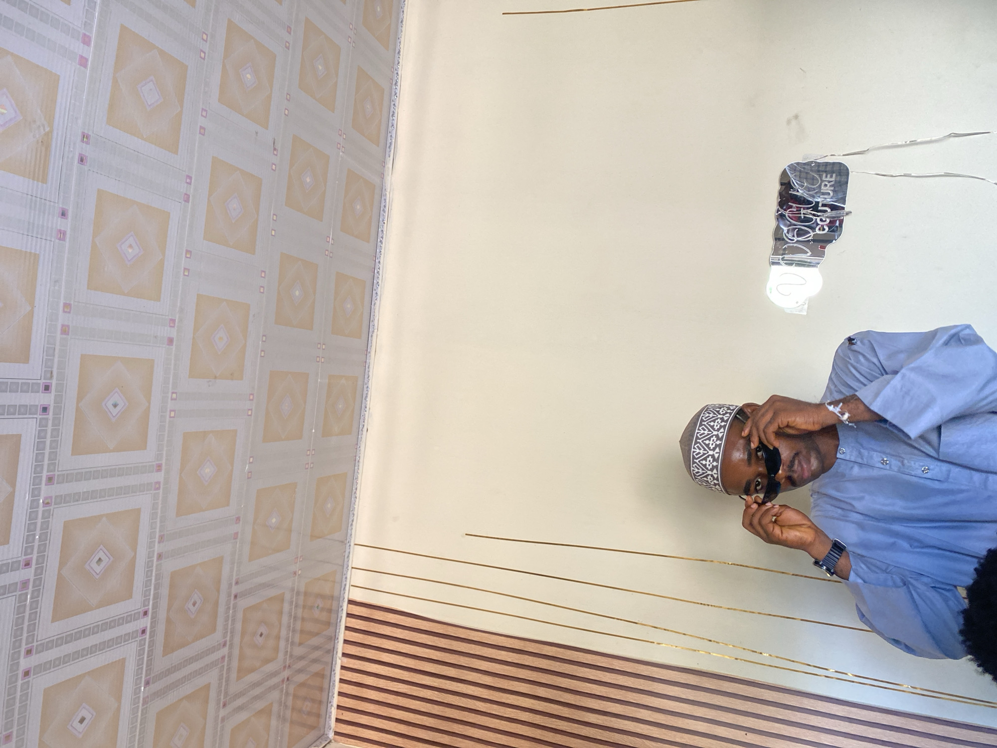 A young man wearing a blue kaftan and a patterned kufi hat is standing indoors, adjusting his black sunglasses. He is looking towards the camera, with his hands on the temples of the sunglasses. To his left, a wall is decorated with vertical wooden slats and thin gold lines. To his right, a neon sign that reads "VOGUE COUTURE" is attached to the wall. The ceiling is a patterned white and beige tile. The location appears to be Jalingo, Nigeria.