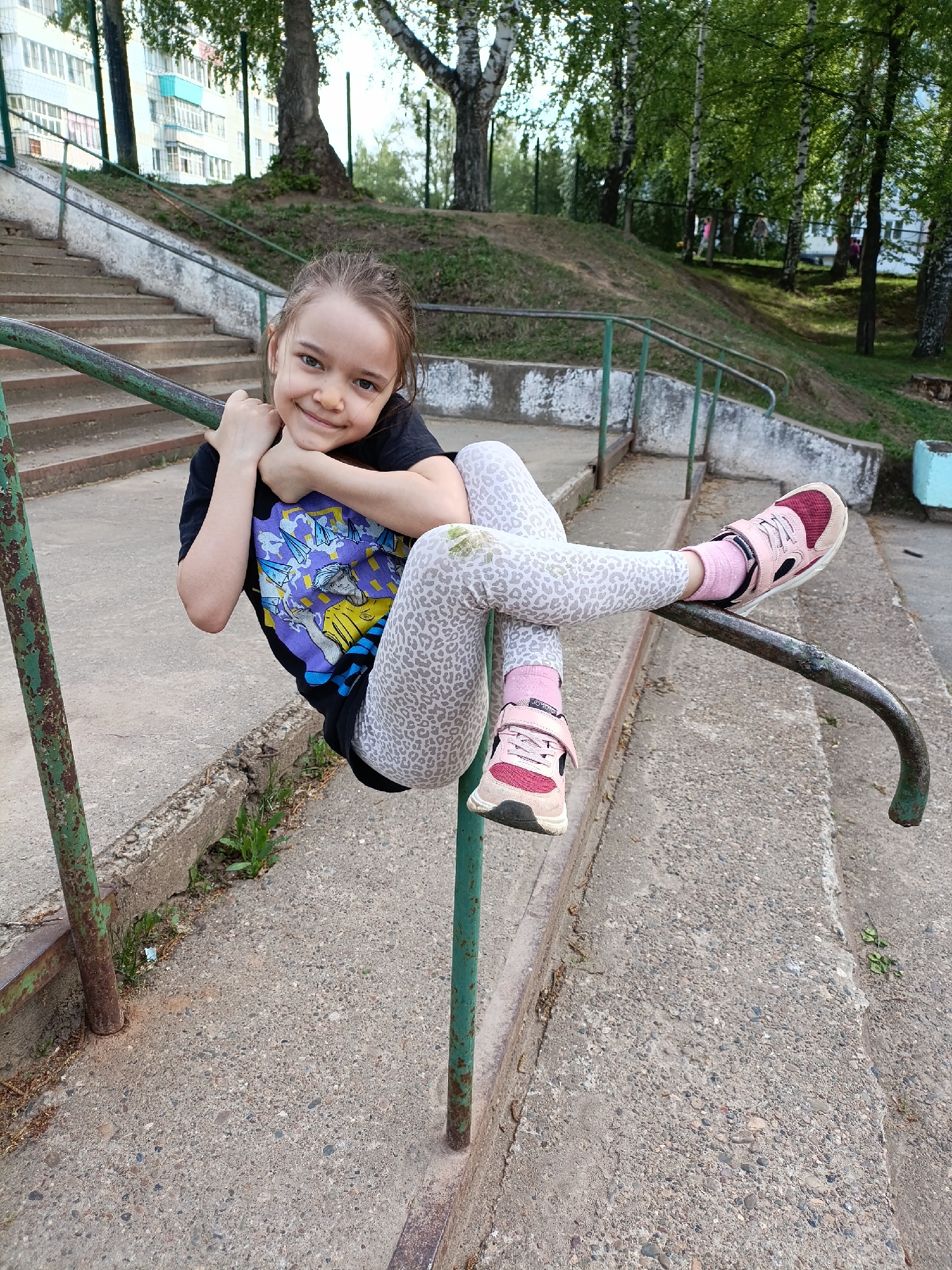 A young girl is suspended from a green metal handrail on a set of concrete stairs. She is wearing a black t-shirt with a colorful graphic design, light gray patterned leggings, pink socks, and pink sneakers. She is smiling and looking directly at the camera. The background includes a grassy hill, trees, and a multi-story building. The location appears to be Chaikovsky, Russia.