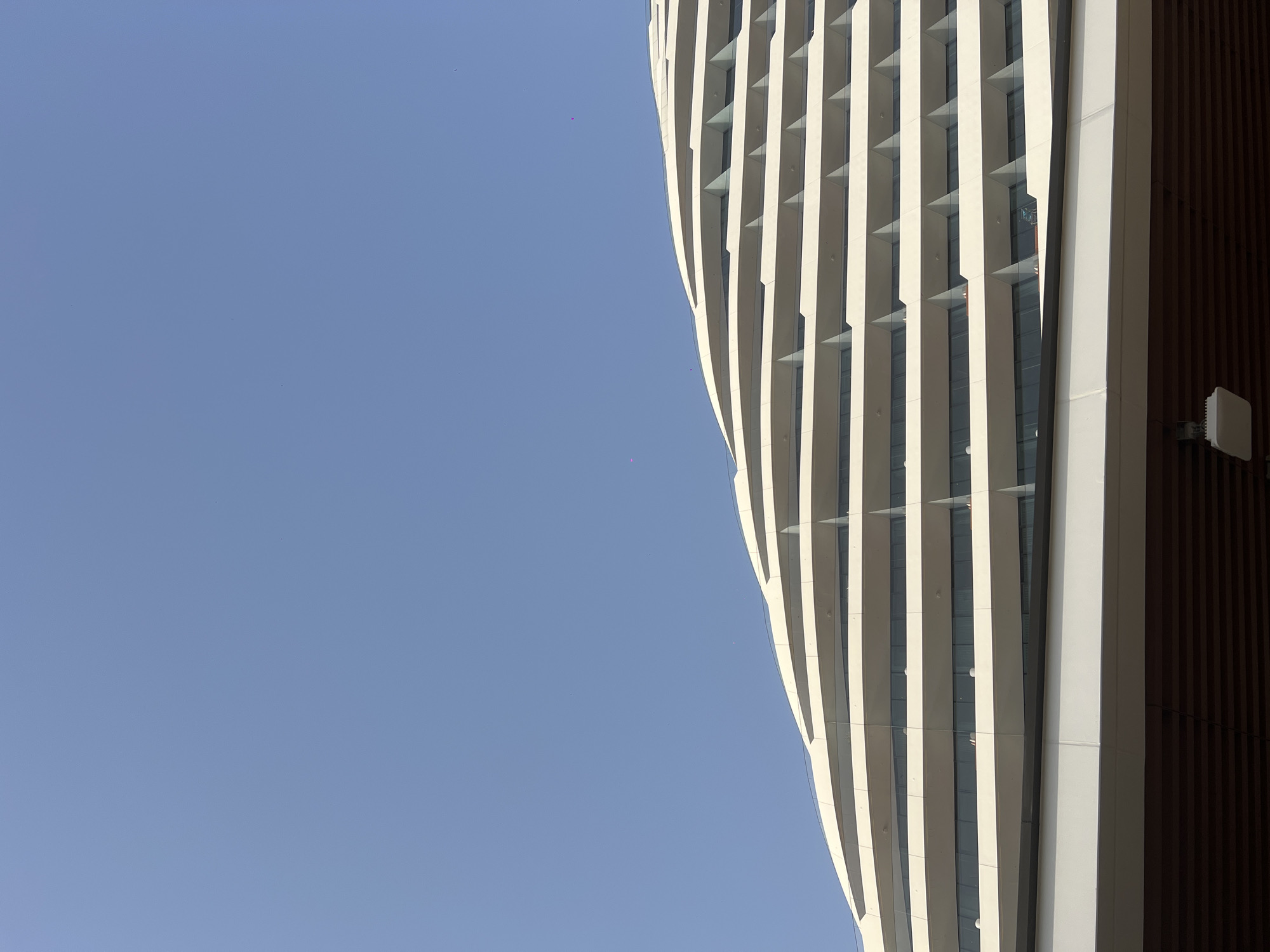 A low-angle shot of a modern building against a clear blue sky. The building has a unique design with white, vertical stripes that create a curved effect. The right side of the building is covered in brown panels. A small, square object, possibly a sensor or antenna, is attached to the brown paneling. The location is Dubai, United Arab Emirates.
