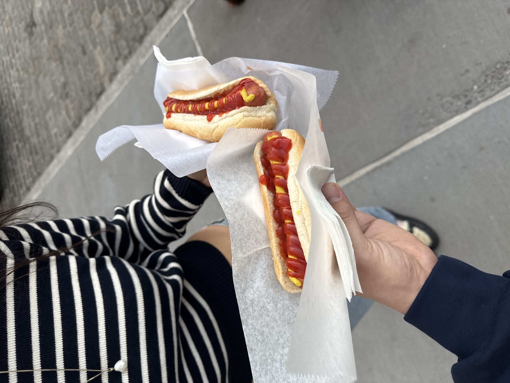 The image captures two hot dogs held in hand, with one person wearing a striped sweater and another wearing a blue jacket. Both hot dogs are on paper towels and are topped with mustard and ketchup. The setting appears to be outdoors, possibly in New York City, USA, given the hot dog type commonly associated with the city. The surface in the background is concrete.