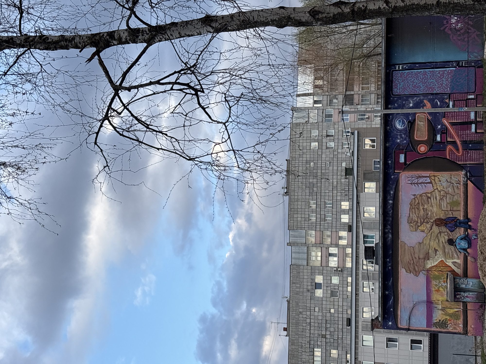 The image captures an urban scene in Perm, Russia, featuring a stark contrast between natural elements and human-made structures, adorned with a vibrant mural. The perspective is tilted, with a large, bare-branched tree in the foreground, silhouetted against a cloudy sky. Its branches, devoid of leaves, extend across the upper left portion of the frame, suggesting late autumn or early spring.
The sky is partly overcast, with varied cloud formations ranging from dark grey to lighter, brighter patches. A faint, diffused sun is visible through the clouds.
To the right, a series of tall, multi-story residential buildings with a typical Soviet-era panel construction dominate the background. These grey buildings have numerous windows, some reflecting the sky.
A significant portion of one building's facade is covered by an elaborate, colorful mural. The mural is divided into distinct sections:
* The uppermost part features abstract, patterned designs in blues and purples.
* Below this, a dark, cosmic-themed section displays stars, a glowing celestial body, and an abstract, capsule-like object with an orange outline and an exclamation mark. Orange glowing lines and shapes extend from it.
* The lowest visible part of the mural depicts a natural landscape with rugged, brown and beige rock formations, sparse trees, and a small body of water or sky at the horizon. Two figures are seated or kneeling within this landscape: one figure with blue clothing is seated, and another with a teal jacket appears to be drawing or painting on a small canvas.
The scene combines urban architecture with natural elements (tree, sky) and a striking piece of public art, suggesting a dynamic city environment. No discernible text is visible in the image.