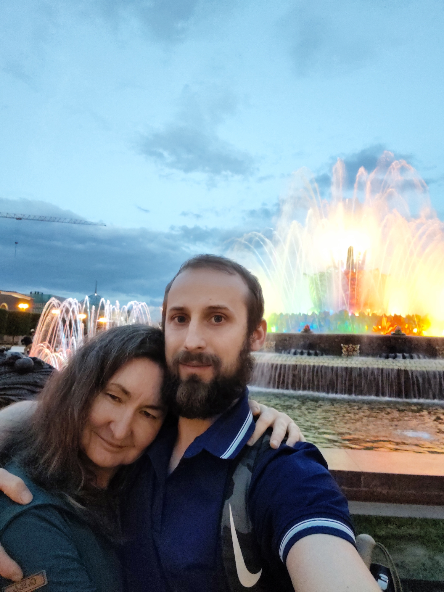 A man and woman pose for a selfie in front of a fountain at the Exhibition of Achievements of National Economy (VDNKh) in Moscow, Russia. The woman, on the left, has shoulder-length brown hair and wears a dark green shirt. Her left eye is partially closed, as if she is winking. The man, on the right, has short brown hair and a beard, and wears a dark blue collared shirt. His left arm is around the woman's shoulders. Behind them, a large, colorful fountain sprays water, illuminated with various colors. In the background, other fountains are also lit. The sky is overcast with a mixture of blue and gray clouds. A construction crane is visible in the upper left of the frame.