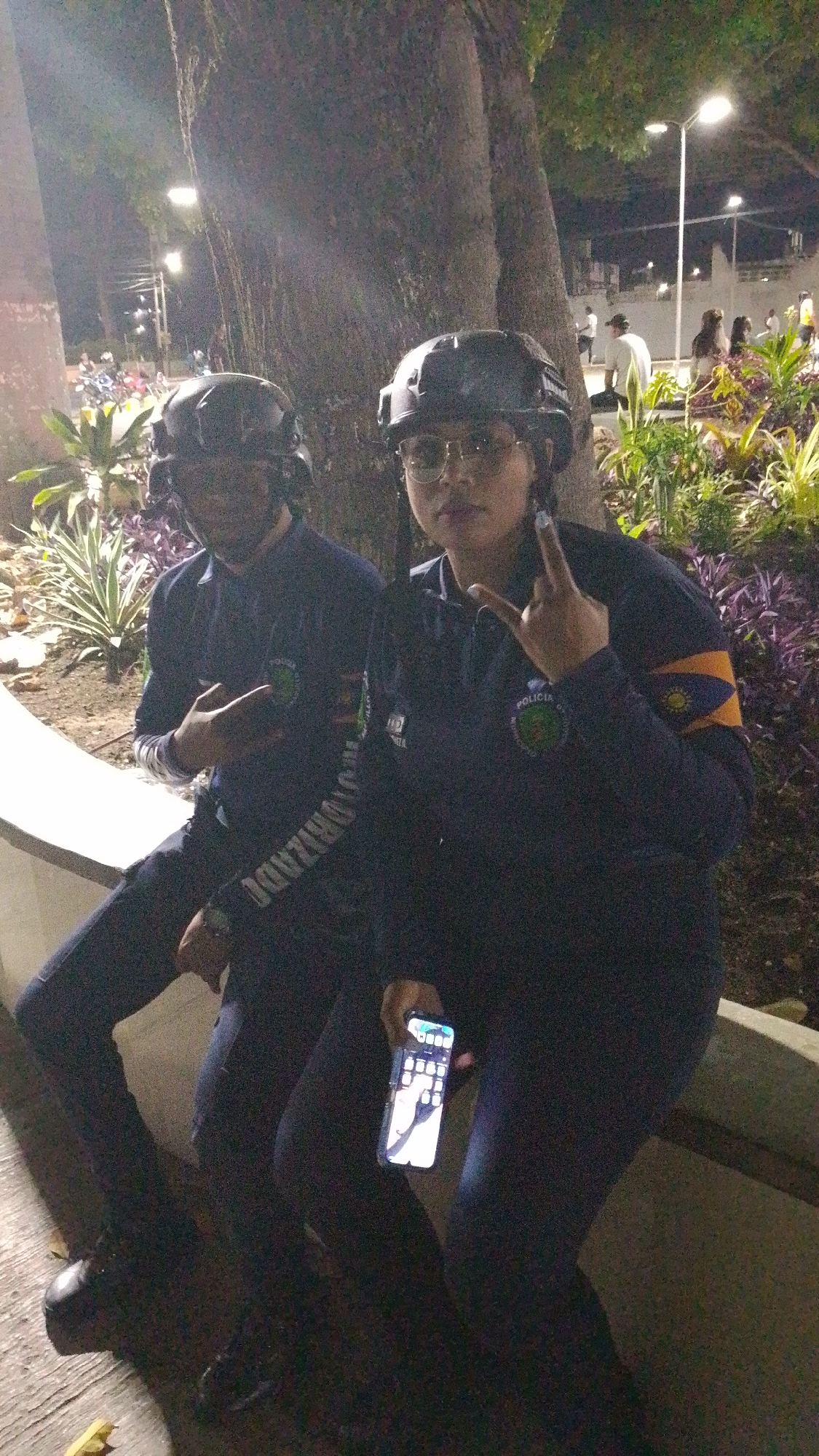 Two people, both wearing helmets and police uniforms, are sitting together outdoors at night. The person on the left is wearing glasses and holding a cell phone. The person on the right is making a hand gesture. They are in the city of Tinaquillo, Venezuela. Streetlights and other people can be seen in the background.