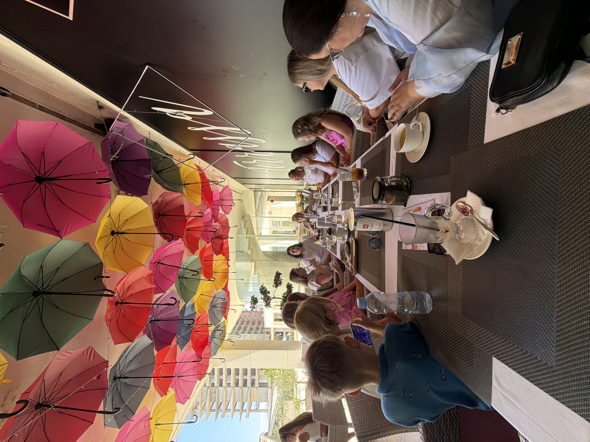 In Budva, Montenegro, a group of people are dining at an outdoor restaurant. The scene is illuminated by natural daylight, casting shadows and highlighting the vibrant colors of numerous umbrellas suspended overhead. These umbrellas, in shades of pink, green, red, yellow, and purple, create a festive canopy above the seating area.
At the foreground, a young boy wearing a blue polo shirt is seated facing away from the camera, engrossed in a mobile device. A water bottle and a partially consumed beverage are placed on the table before him. To his right, a woman with blonde hair and sunglasses is seated, looking towards her left. Further down the table, a woman in a light blue top is actively engaged with her phone, her hands resting on a table. Next to her, another woman, also in a light blue top, is seated.
The table is set with placemats, cutlery, glasses, and plates, indicating a meal is in progress. The background reveals more diners and a glimpse of the restaurant's interior, characterized by modern architecture and glass elements. Construction cranes are visible in the distance, suggesting ongoing development in the vicinity. The overall atmosphere suggests a relaxed outdoor dining experience.