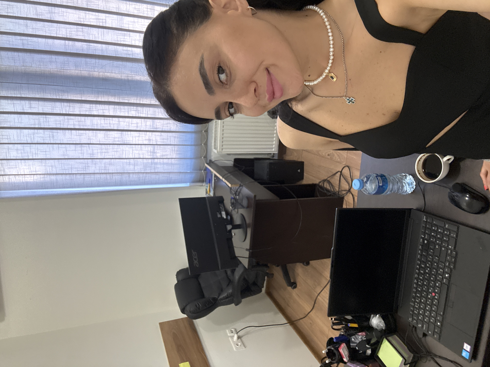 The image depicts a woman in an office setting, likely in Tbilisi, Georgia, based on the context provided. She is seated at a desk, facing the camera with a slight smile. The desk is equipped with a laptop, a computer mouse, a cup of coffee, and a bottle of water. Behind her, an office chair and a computer monitor are visible. The environment appears to be a typical workspace with a window covered by vertical blinds. The woman is wearing a black top and a pearl necklace. The overall impression is a candid snapshot of someone in their workplace.