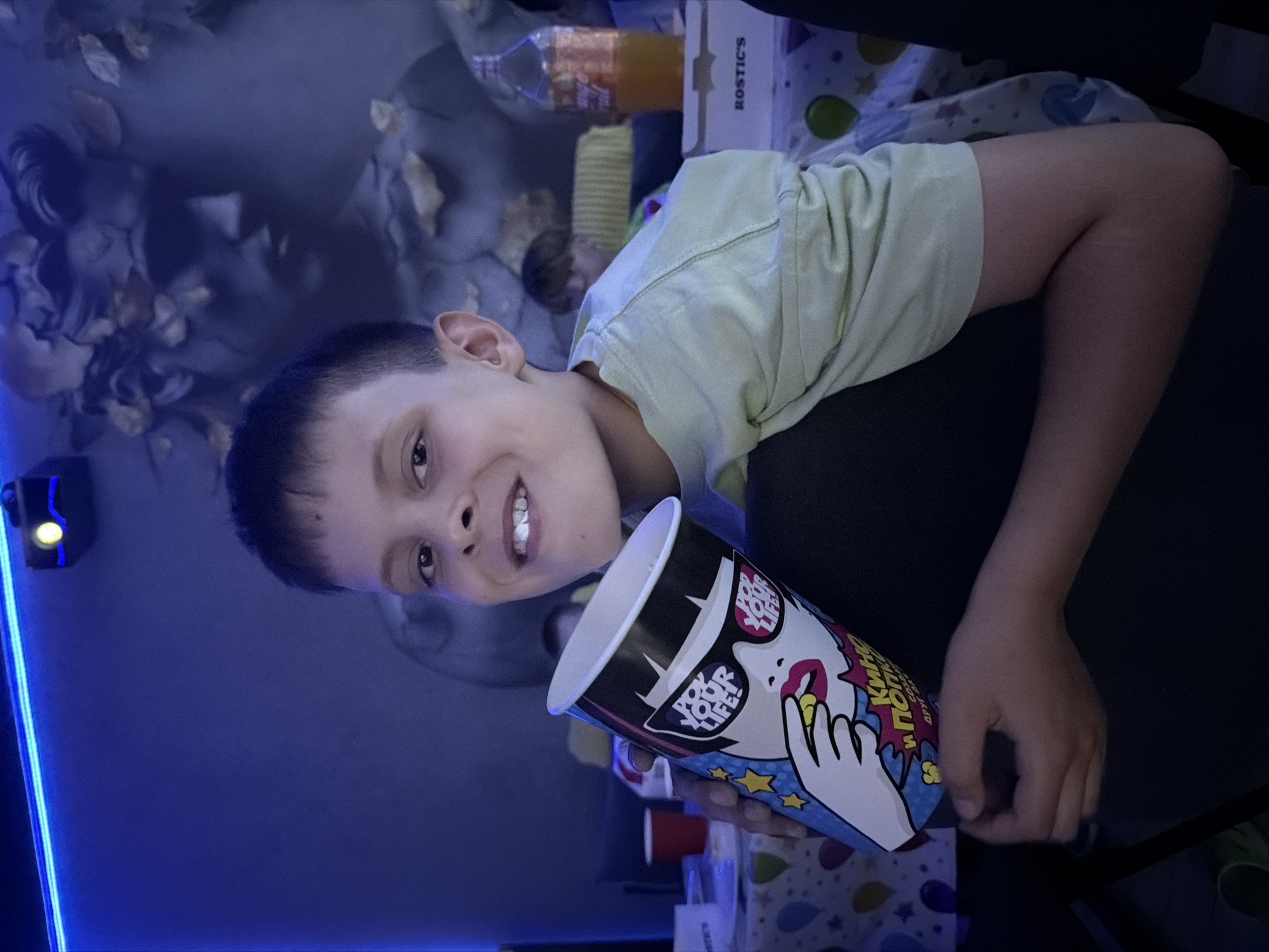 A young boy, appearing to be between 8 and 10 years old, smiles at the camera while holding a large popcorn container. He has short, dark hair and is wearing a light green t-shirt. The popcorn container has a colorful, cartoonish design featuring a woman with sunglasses and the text "POP YOUR LIFE!". He is holding a piece of popcorn to his mouth. The boy is seated, with his arm resting on his leg. In the background, there is a dark wall with a large, dark gray statue of a face with leaves on its head. A blue light strip runs along the top of the wall, and a projector is mounted near it. To the right, there is a bottle of orange liquid and a white box with the text "ROSTIC'S". A table covered with a patterned tablecloth is visible, with red cups on it. The overall lighting is dim and predominantly blue, suggesting an indoor environment, possibly a home theater or a private viewing area, set up for a movie or event. The image does not provide any specific visual cues to pinpoint its location within the city of Votkinsk, Russia.