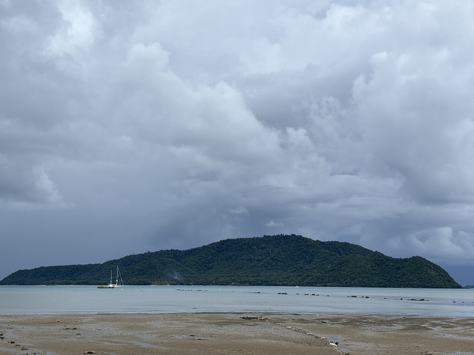 The image shows a wide shot of a beach in Rawai, Thailand. In the foreground, a sandy beach with scattered debris and some shallow water is visible. A calm sea stretches out towards a lush, green, tree-covered island in the mid-ground. The island is elongated and curves across the horizon. Several boats are present on the water: a prominent white sailboat with two masts is anchored in the middle distance, and other smaller boats or buoys are scattered further out. The sky is overcast with dramatic, heavy clouds, suggesting an impending storm or recent rain. The overall scene is natural and coastal.