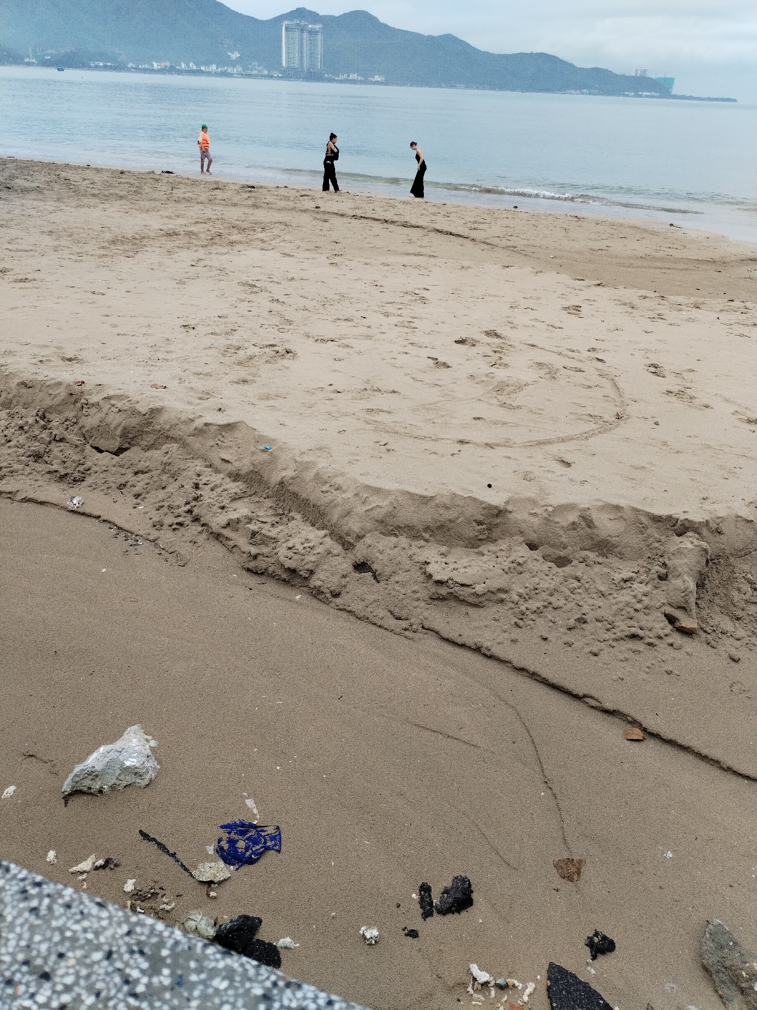 The image captures a coastal scene at Bac Nha Trang Ward, Vietnam, under an overcast sky. In the midground, three people are visible near the calm ocean. To the left, an individual in an orange top and grey pants wades in the shallow water. Two other people, both dressed in dark clothing, walk along the waterline further to the right.
The sandy beach dominates the foreground, featuring a prominent sand erosion line that forms a small, irregular cliff across its mid-section. Above this erosion, faint circular patterns are drawn into the sand. In the immediate foreground, scattered debris and litter, including plastic pieces and other small items, are visible on the sand. A speckled concrete structure partially occupies the bottom left corner.
The background shows the expansive ocean meeting a distant coastline, marked by a range of rolling mountains and several modern high-rise buildings, indicating a developed urban area by the sea. The overcast sky provides soft, diffused lighting across the entire scene. The atmosphere suggests a quiet, cloudy day at the beach, characterized by human activity near the water and scattered litter on the sand. No discernible text is visible in the image.