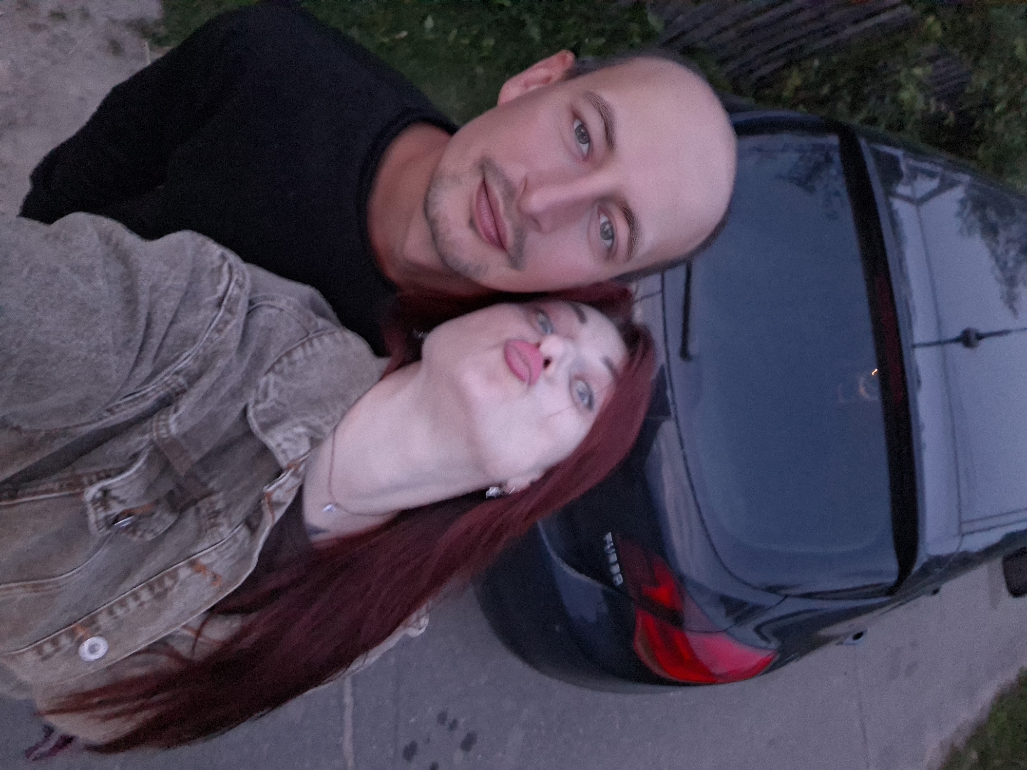 A selfie is taken by a couple in front of a dark-colored hatchback car on a paved road, with greenery visible to the left and right. The man, on the left, has a shaved head and is wearing a black t-shirt. He is looking towards the camera with a neutral expression. The woman, on the right, has long, dark red hair and is wearing a denim jacket. She is puckering her lips and looking slightly upwards. The background shows the rear of the car and a hint of trees and a fence. The overall scene suggests a casual outdoor setting, possibly a rural road or the outskirts of a village. The quality of the image is somewhat grainy, suggesting it might have been taken in low light conditions, possibly at dusk or dawn. There is no specific indication of it being in Забалоцкі сельскі Савет, Belarus, but the description is based on the provided image.