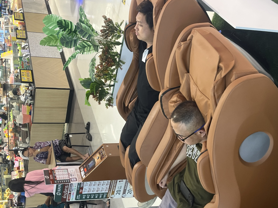 The image displays an indoor commercial environment in Bang Pu Subdistrict Municipality, Thailand, consistent with a mall food court or supermarket section. In the foreground, two individuals are reclined in tan-colored full-body massage chairs, situated on a green turf-like carpet. The individual closest to the viewer is a male with a receding hairline and eyeglasses, wearing a dark green shirt, with his head resting in the chair's headrest. Adjacent to him, a second male with dark hair and a black shirt is similarly reclined in an identical massage chair.
To the left of the massage chairs, a brown payment kiosk or control panel for the chairs stands upright, featuring a digital display and a vertical sign with pricing information predominantly in Thai, with "KAYA" visible. To the right of the chairs, a white rectangular planter holds a variety of artificial plants, including large green striped leaves and smaller reddish-brown foliage.
In the mid-ground, a brightly lit food service area is visible, characterized by display counters with glass cases containing various food items. A woman wearing a pink top stands near a small, round, light-colored table. An older male with eyeglasses, dressed in a patterned short-sleeved shirt and dark shorts, sits barefoot at this table, with dark-colored sandals resting on the light-colored floor beside his chair. He appears to be observing the area or the woman. Further in the background, additional individuals, some appearing to be staff members wearing face masks, are present behind the food counters. The floor throughout the visible area is a clean, reflective light-colored surface.