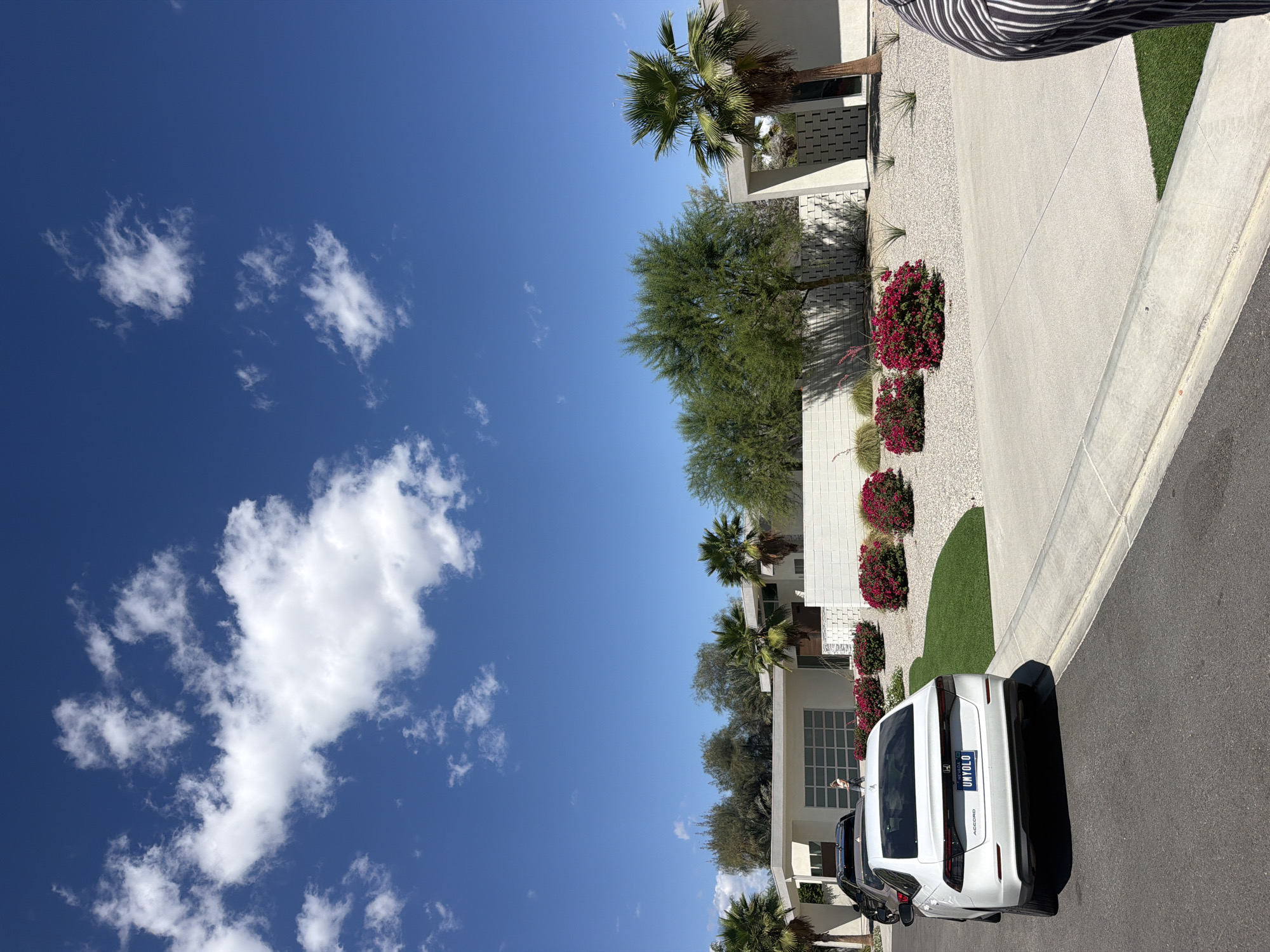The image captures a sunny day in what appears to be a residential area, possibly Indio, California, given the desert landscaping, architecture, and license plate. The sky is predominantly blue, dotted with fluffy white clouds. A modern, white single-story building with palm trees and desert vegetation is visible. Along the driveway, there are red flowering bushes and gravel landscaping. A white car is parked on the street, its license plate partly obscured, reading "UNYOLO". A person stands near the car. The focus is on the interplay of architectural design, landscaping, and a clear sky.