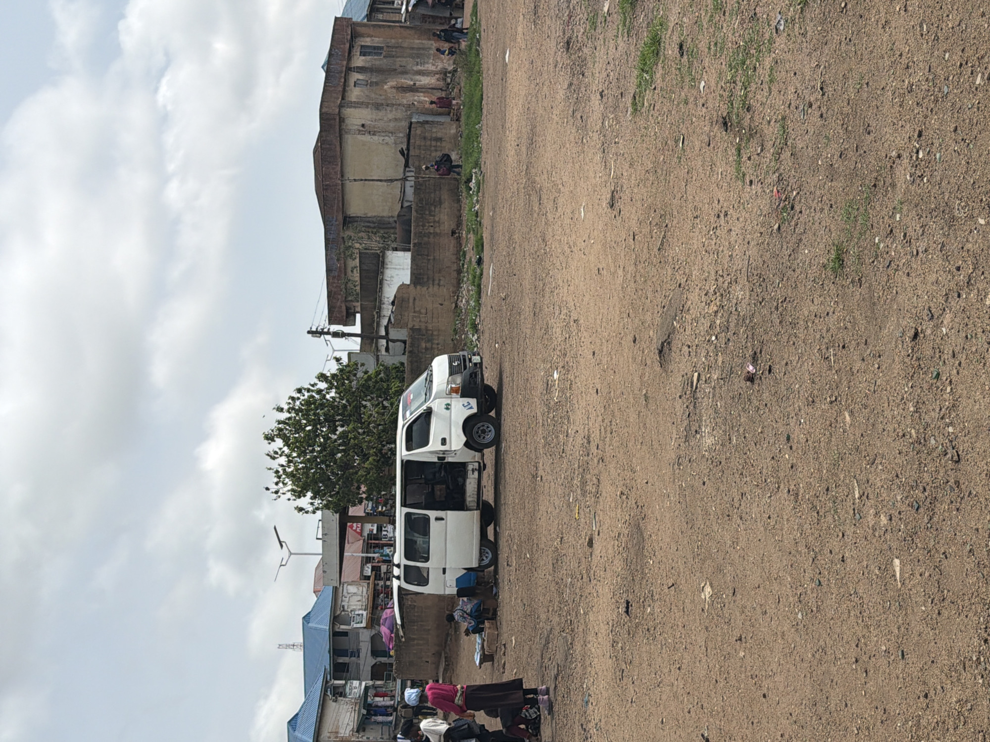 This image depicts a street scene in Bukuru, Nigeria. In the foreground, a dirt and gravel expanse is sparsely scattered with debris. To the left, a white van with its side door open is parked. Near the van, several individuals are present, including one person wearing a pink shirt and a headscarf, and another person sitting on the ground. In the background, there are buildings with blue and red roofs, a tree, and a brick wall. Further in the background, more structures are visible, including a large, older building with a brown roof. The sky is partly cloudy.