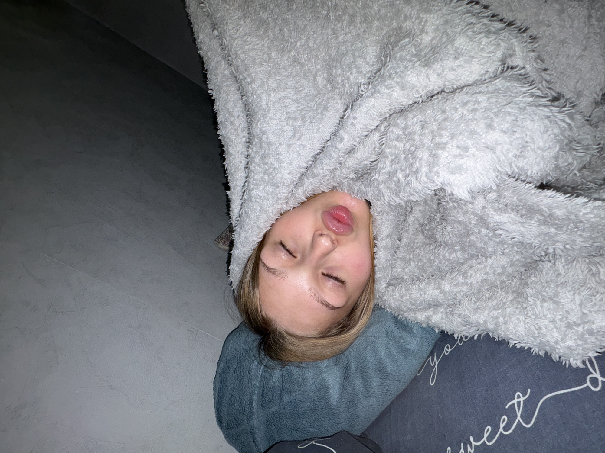An overhead, rotated photograph captures a young person seemingly asleep or deeply resting indoors. The individual, whose face is visible, is covered largely by a light gray, plush, and fluffy blanket or hooded garment. Their eyes are gently closed, and their lips are slightly parted, suggesting a state of peaceful slumber. The person's head, with light brown hair, is supported by a solid blue-teal pillow. Beneath this, a darker blue-gray pillow is partially visible, featuring white cursive text that reads "you" and "sweet," likely part of an affectionate phrase. The background consists of a plain, textured gray surface, possibly a wall or floor. The lighting in the scene is dim, indicating it could be nighttime or a softly lit room. The overall impression is one of calm and relaxation. The image itself provides no direct geographical cues, but the context provided is Chaikovsky, Russia.