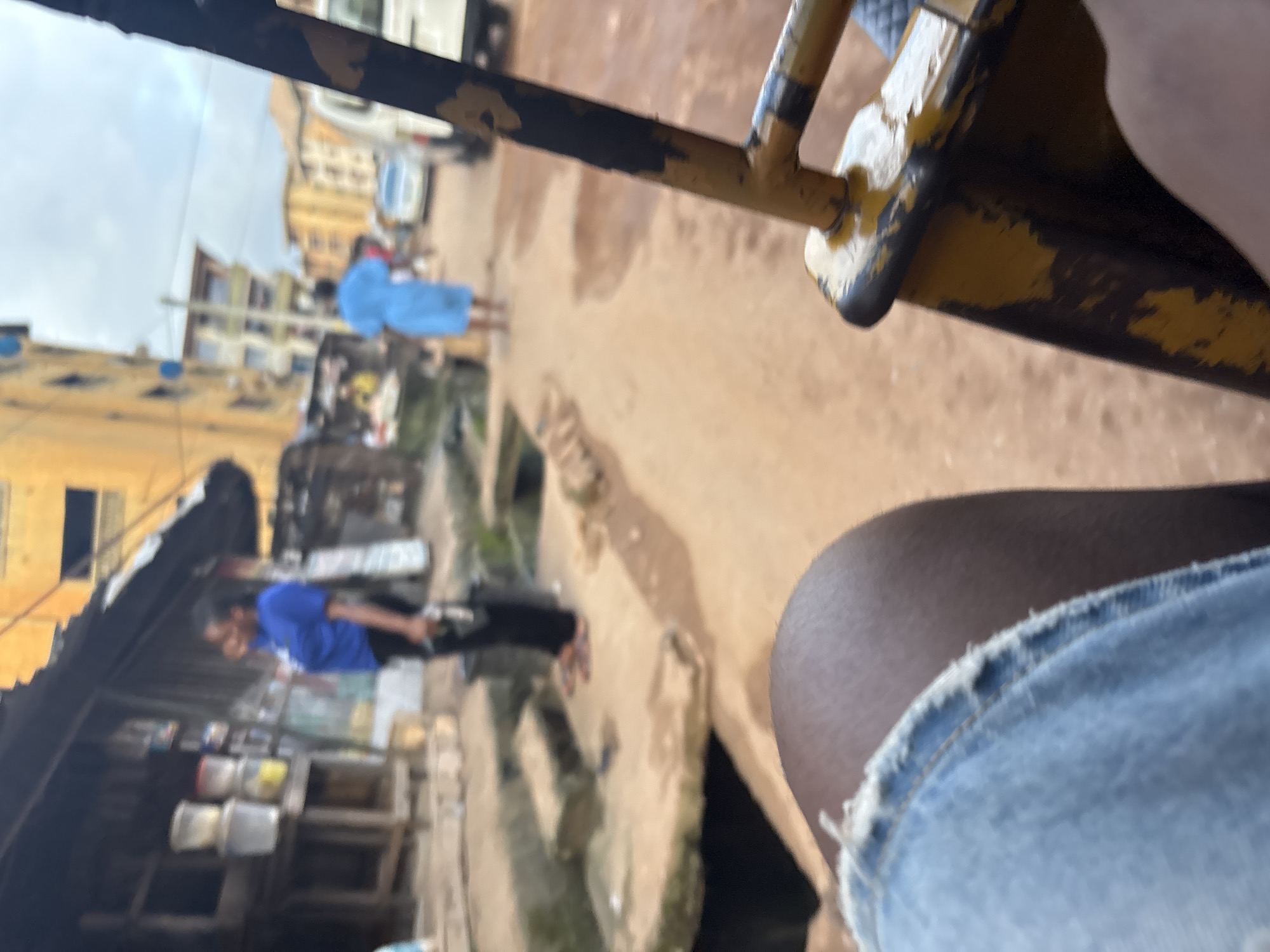 The image depicts a street scene in Onitsha, Nigeria, likely from the perspective of someone seated in a vehicle, possibly a motorized rickshaw. In the foreground, a person's knee and thigh, clad in denim shorts, are visible. The environment is characterized by a dusty, reddish-brown unpaved road.
To the left of the frame, a small, open-fronted shop or stall is visible. It has a corrugated metal roof extending outwards. On shelves within the stall, several plastic containers or buckets are stacked. A woman wearing a bright blue t-shirt and dark trousers stands in front of this stall, holding a dark object in her right hand. Her body is angled away from the camera, and her left arm is bent.
Further down the street, to the right, another individual is seen wearing a light blue garment, possibly a dress or robe, and is facing away from the camera. A white vehicle, which appears to be a minivan or small bus, is parked or moving in the background, alongside other buildings. The buildings in the background are multi-story, with some exhibiting a yellowish facade. The sky is overcast. A metal frame, painted yellow and black with visible peeling paint, occupies the right side of the foreground, suggesting the vantage point is from within a vehicle's structure. The overall impression is that of a bustling, typical urban street in a Nigerian city.