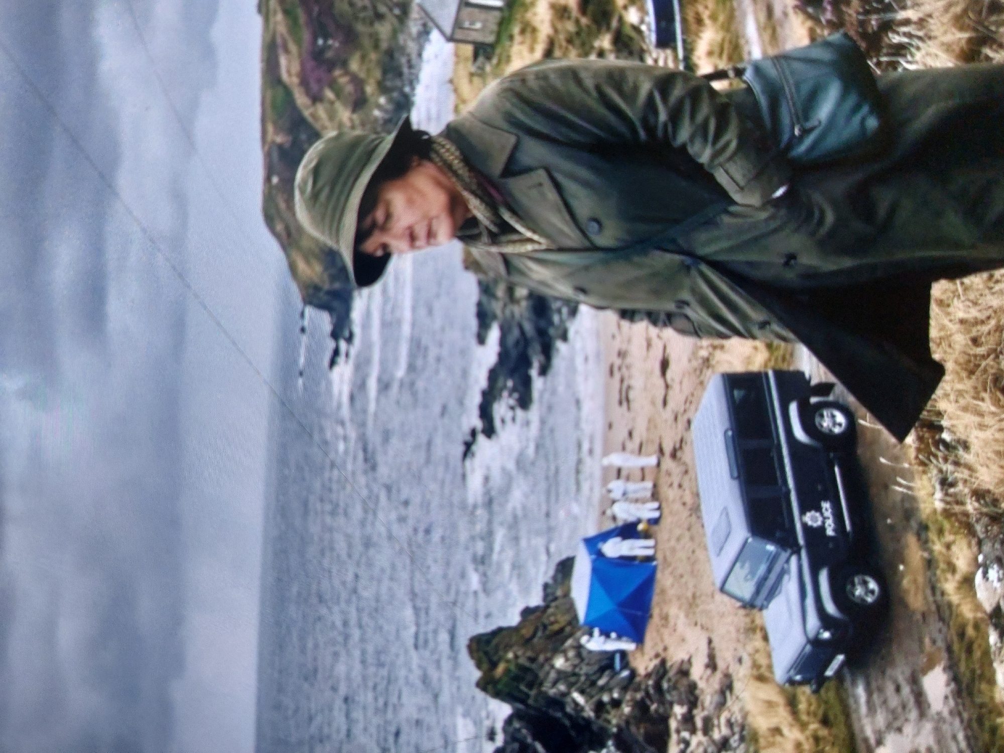 A coastal scene depicts a male figure in a green overcoat and hat standing near a rocky shoreline. In the background, a vehicle marked "POLICE" is parked near a blue tent, with figures in white suits present on the beach. The scene evokes a potential forensic investigation on a beach, but the location is not Tsarevo, Bulgaria. The location is unidentified, but based on similar images, this appears to be the location for the series 'Shetland', filmed in Scotland. The scene involves potential crime scene investigation.