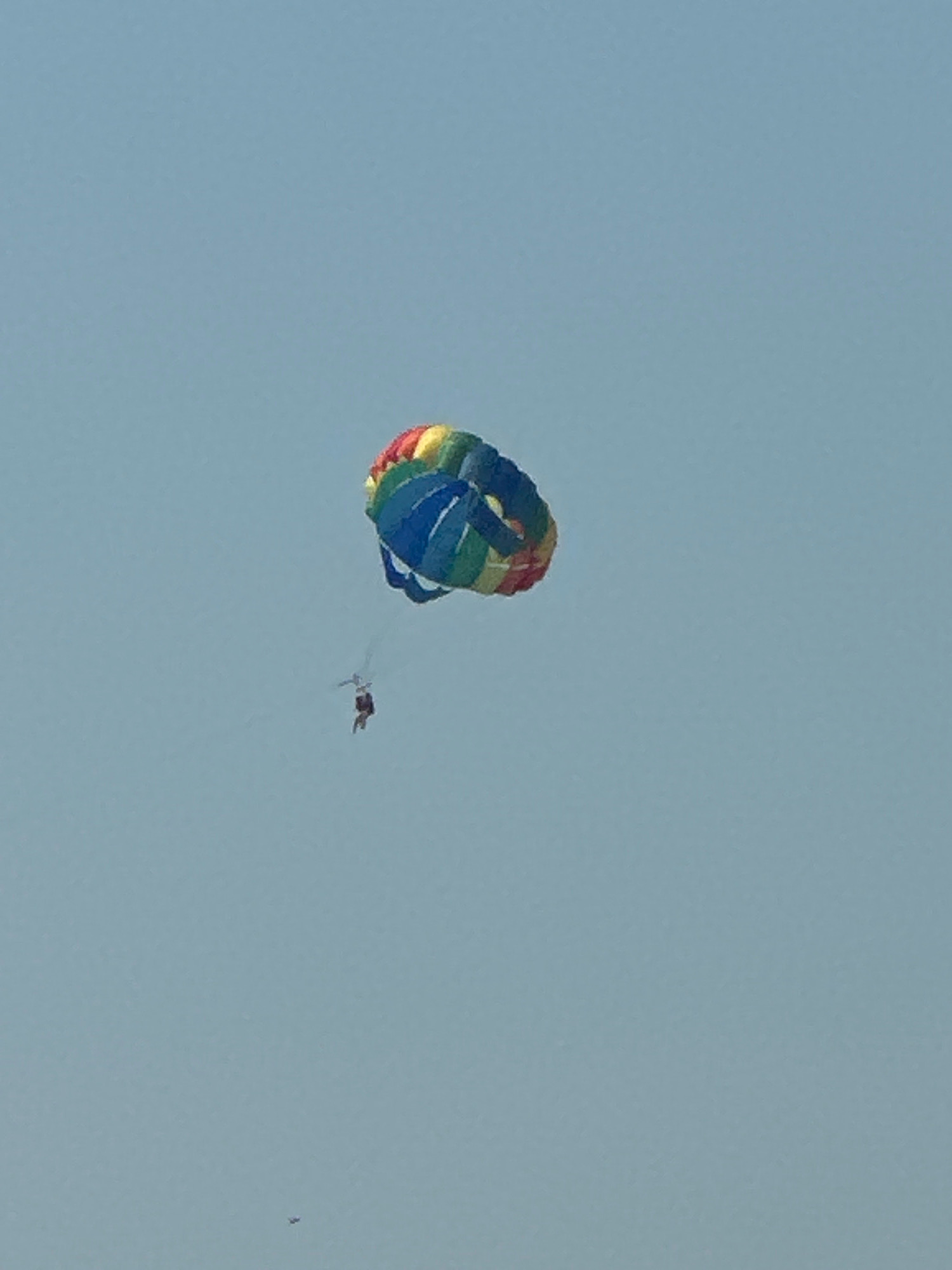 A person is parasailing in the sky over Budva, Montenegro. The parachute is colorful, with stripes of blue, green, yellow, and red. The person is suspended below the parachute by a harness and is wearing a red top and dark shorts. The sky is a clear, pale blue. There is a tiny speck of what might be a bird or a small airplane in the lower left corner of the image.