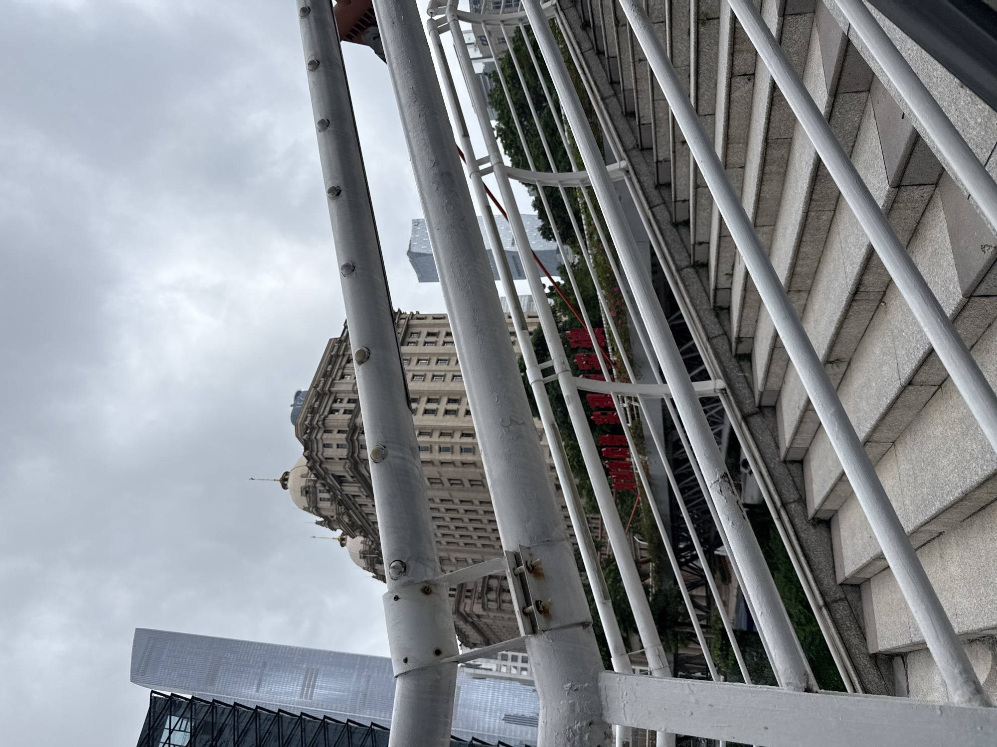 An exterior, eye-level shot shows a white metal staircase and railing in the foreground, with a city skyline in the background under a cloudy sky. The staircase has granite steps. Behind the railing, a row of red banners with white lettering is visible. Further back, an ornate, old building with multiple domes and an arched facade stands to the right, and to its left, a modern, tall glass skyscraper. Additional modern buildings are visible in the distance. The scene appears to be in the Tianhe District of Guangzhou, China, a bustling urban area known for its mix of historical and contemporary architecture. There are no people or animals clearly visible in the image.