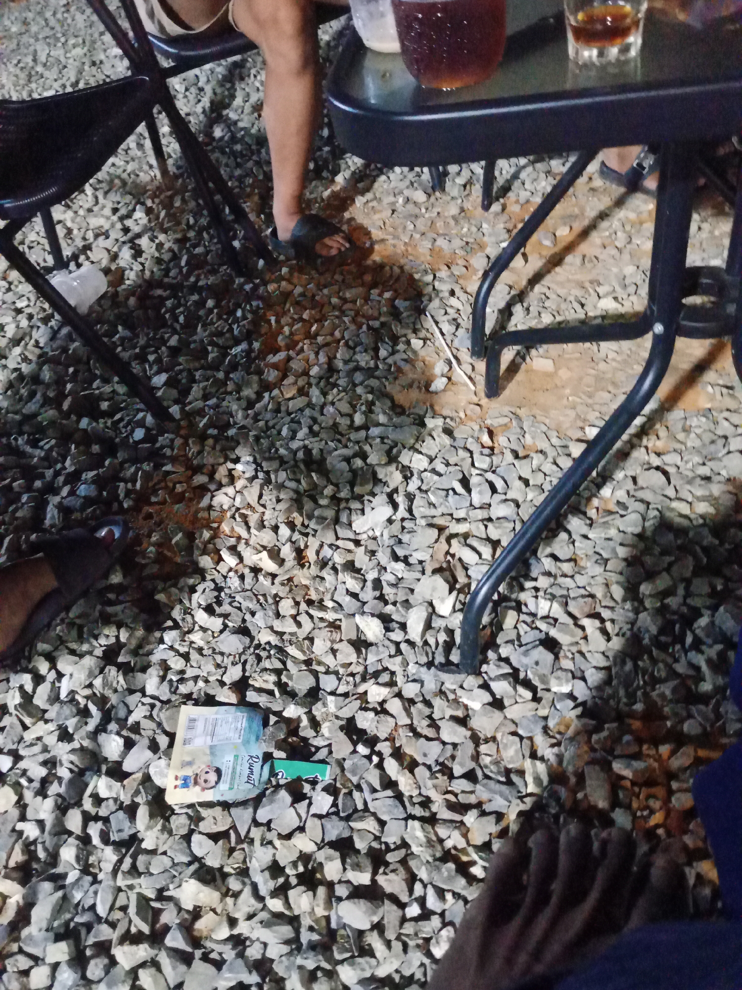 The image depicts an outdoor scene at night on a ground covered with small grey and brown gravel. In the foreground, a person's bare feet are visible, with toes pointing upwards. To the left, a person's legs are seen wearing dark flip-flops, with one foot resting on the gravel. A portion of a black folding chair is also visible on the left. In the mid-ground, a small white package with a cartoon character and text in an Asian language is lying on the gravel. Behind this, a dark metal table with black legs is set up, with a glass containing a dark beverage and another smaller glass with amber liquid on its surface. The overall environment appears to be a casual outdoor gathering space, possibly at a restaurant or bar. The location is identified as Mekar Jaya, Indonesia.