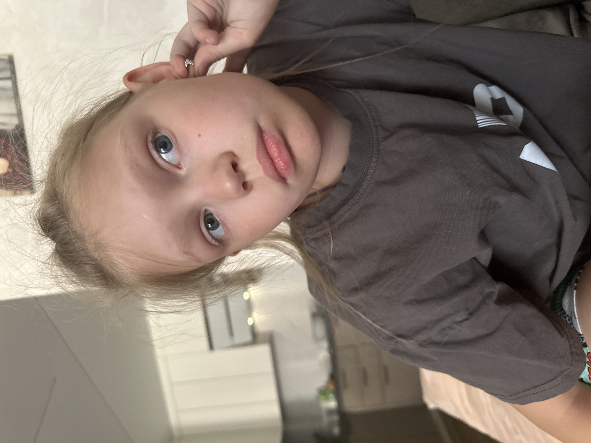 A young girl with blonde hair pulled back is looking to her right with a slightly pouty expression. She is touching her ear, where she is wearing a small, silver earring. She is wearing a dark grey t-shirt. The background shows a blurred kitchen area with white cabinets and kitchen appliances. The image appears to be a close-up indoor shot. There is no indication that this image is located in Abakan, Russia.