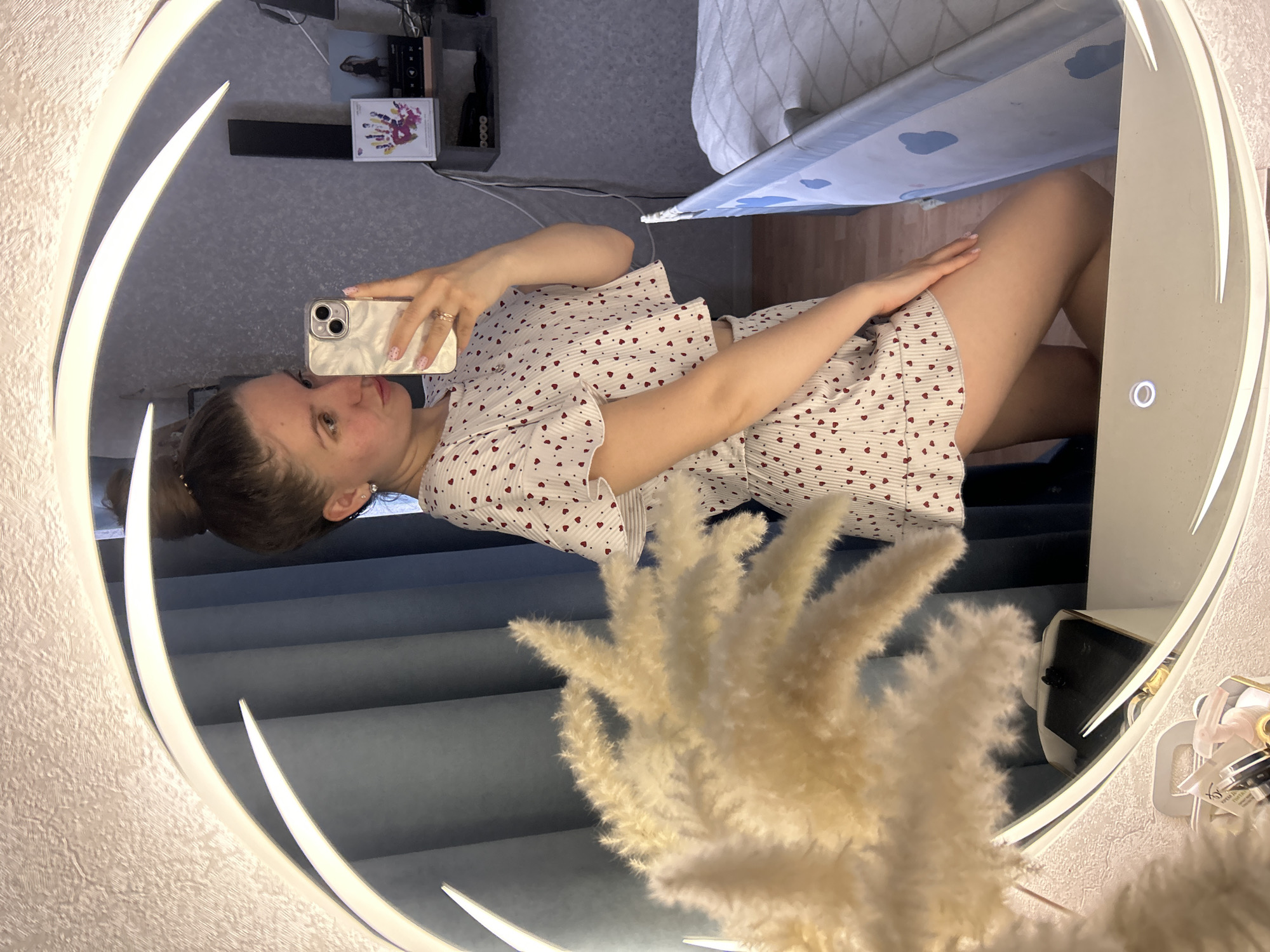 A young woman is reflected in a circular mirror, holding a white mobile phone in her left hand and taking a selfie. She is wearing a white pajama set with small red heart patterns. Her hair is pulled back in a bun. Behind her is a blue wall and a dried floral arrangement partially obscures the reflection. Above and to the side of the woman, in the mirror, is a shelf with various items, including a book with visible handprints. The location is implied to be Klin, Russia. A portion of a bed is visible at the top of the mirror.