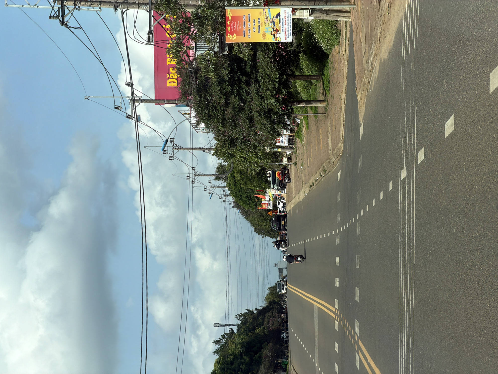 The image depicts a road in Măng Đen, Vietnam, characterized by a gray asphalt surface marked with double yellow lines down the center and dashed white lines delineating lanes. Several vehicles, including cars and motorcycles, are visible on the road. Along the right side of the road is a sidewalk, beyond which are trees and a large red and yellow billboard advertising local information in Vietnamese. Power lines and utility poles stretch across the sky on the left, adding vertical elements to the composition. The sky is partly cloudy.