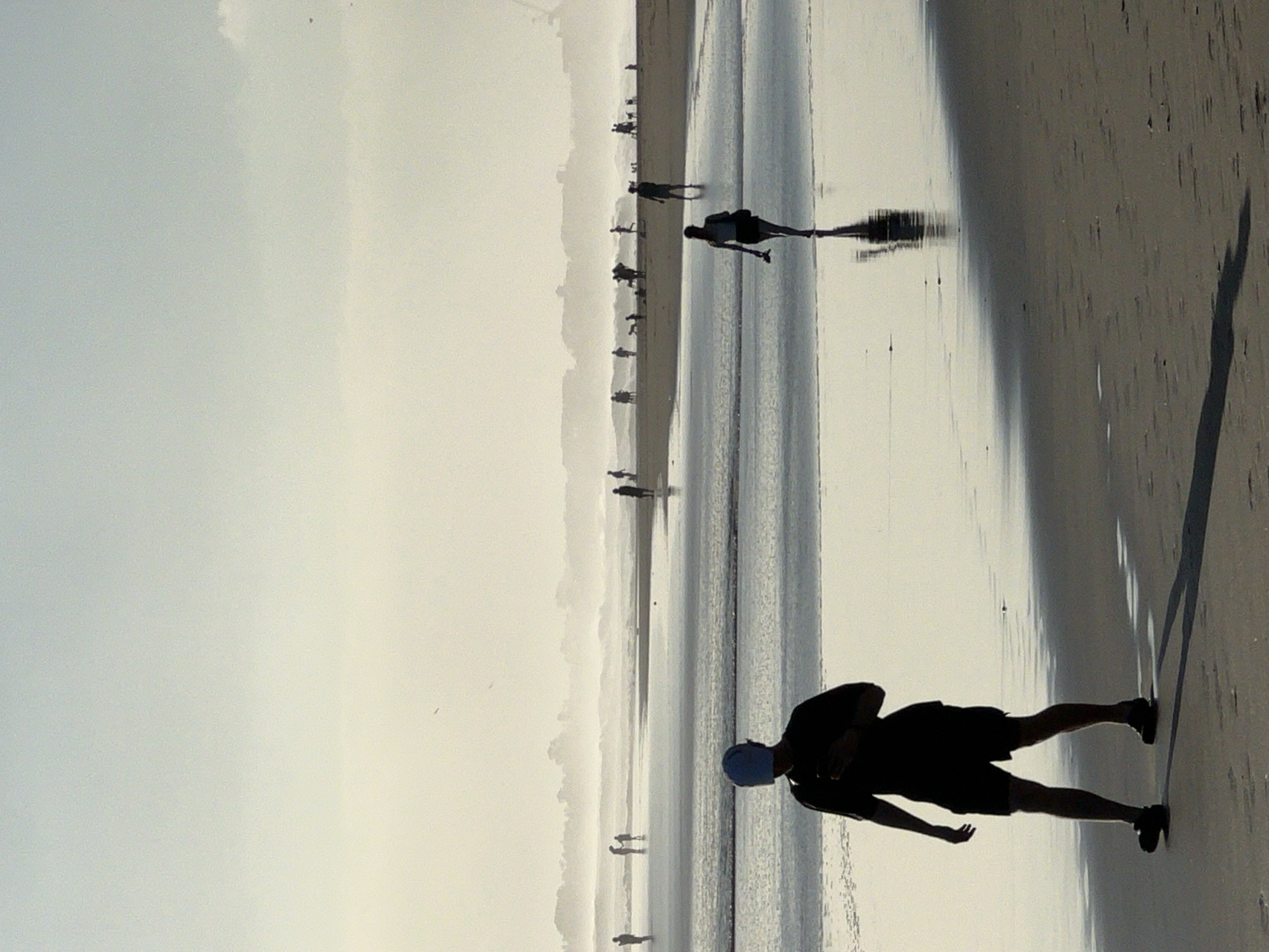 This media file depicts a group of silhouetted figures walking along a beach in North Kuta, Indonesia. The scene is characterized by a wide, flat expanse of wet sand, reflecting the overcast sky. The overall lighting suggests it is either early morning or late afternoon, with a diffused and hazy quality to the light.
The primary subjects are the people, each appearing as a dark silhouette against the brighter background. They are scattered across the beach, with some individuals walking alone and others in small groups. Their postures suggest casual movement, with no specific activity or event being clearly identifiable beyond walking.
The setting is clearly a beach, indicated by the wet sand, the presence of water reflecting the sky, and the distant hazy outline of what might be sand dunes or low hills. The water appears calm, with gentle ripples visible. The sky is a pale, washed-out grey, contributing to the overall subdued and somewhat ethereal atmosphere.
Notable details include the strong, elongated shadows cast by the figures, indicating a low sun angle. The wet sand creates a mirror-like surface in places, further enhancing the reflections. The distant figures are less distinct, appearing as small specks against the horizon, emphasizing the vastness of the beach. There is no visible text in the media file. The overall mood is peaceful and contemplative, with the silhouettes suggesting a sense of anonymity and universality.