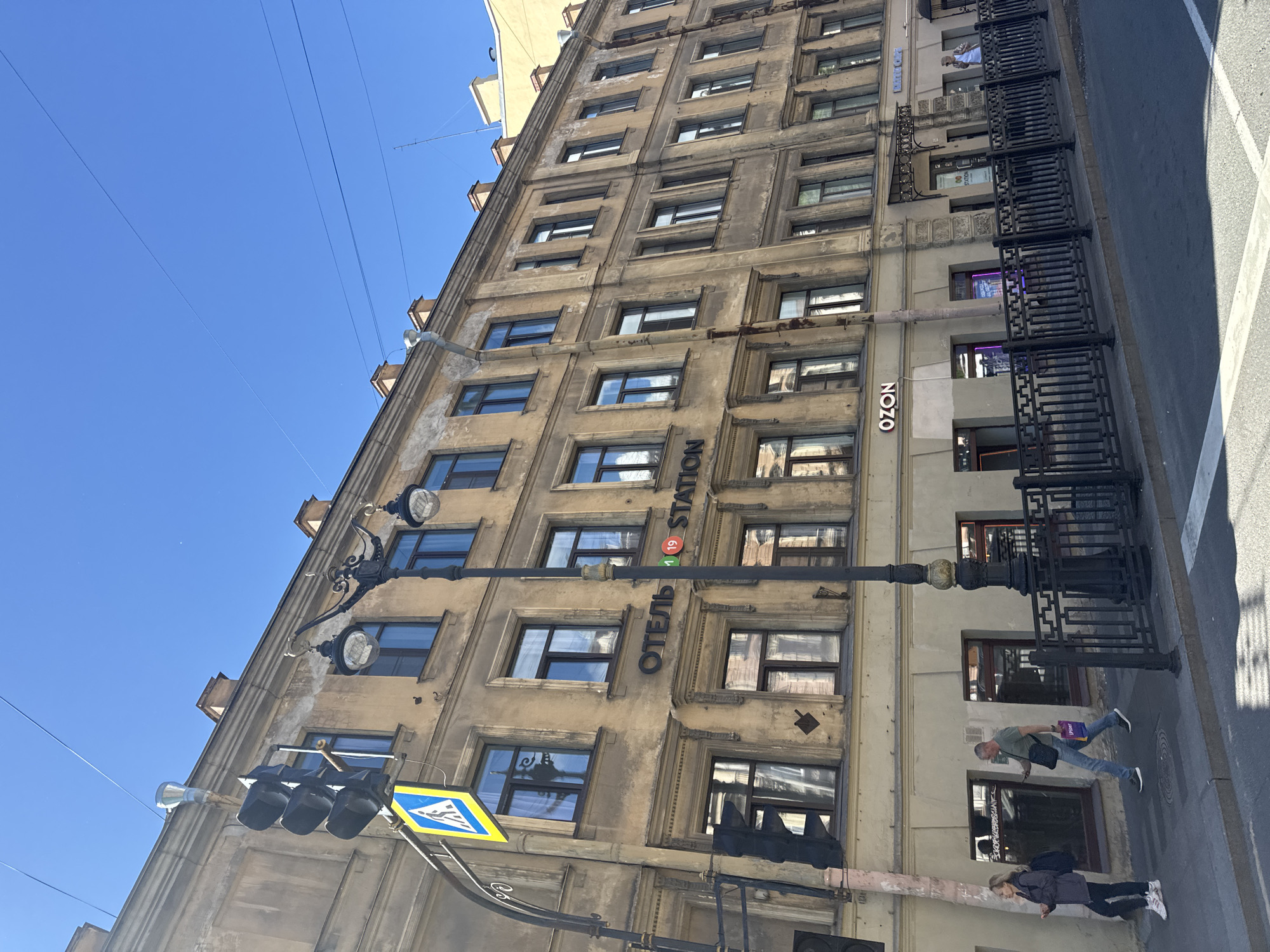 The image captures a street scene in Saint Petersburg, Russia, on a bright, sunny day. A tall, old building with numerous windows and a weathered facade dominates the frame. The building appears to be a hotel, as indicated by the Cyrillic text "ОТЕЛЬ STATION" (Hotel Station) visible on its side, accompanied by a red circle with the number "19". Below the hotel sign, the logo "OZON" is also visible, suggesting a retail presence.
In the foreground, a traffic light with three black lamps hangs above the street. A yellow rectangular pedestrian crossing sign with a blue border is also visible, indicating a crosswalk. The street itself is paved, and a black metal fence runs along the sidewalk.
Two people are seen walking on the sidewalk. One, a man in a green t-shirt and jeans, is carrying a purple bag. He is walking away from the camera. The other person, a woman in a dark jacket and jeans, is walking towards the camera, also with a bag.
The clear blue sky and the bright sunlight suggest it is daytime. The architectural style of the building and the presence of Cyrillic text strongly point to the location being Saint Petersburg, Russia. The overall scene depicts a typical urban environment with people going about their day.