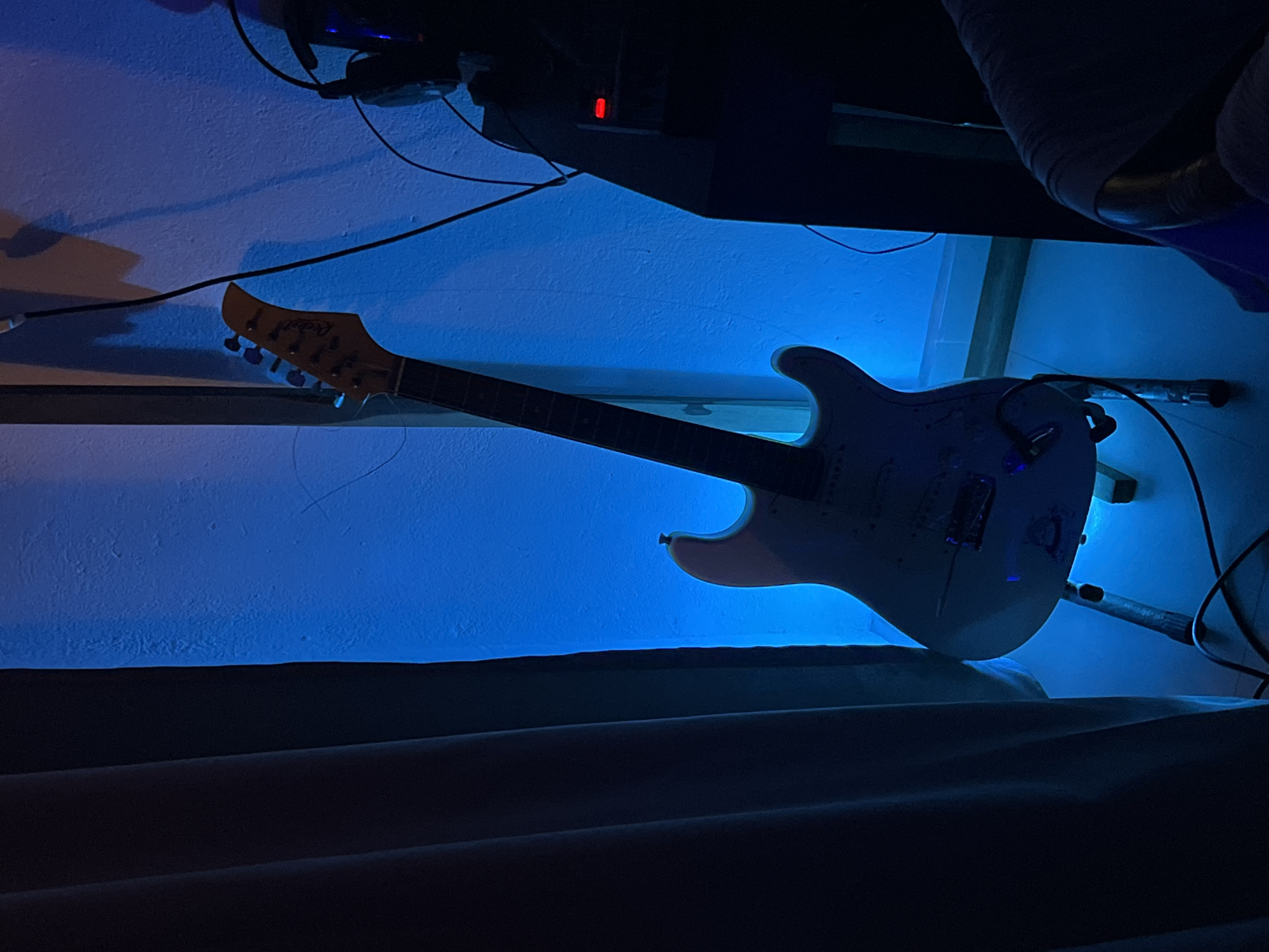 A white electric guitar hangs on a wall. The wall is painted blue and illuminated by a blue light source. Electrical wires and a black box are mounted above the guitar. A dark, soft material covers the bottom of the image. The location is unknown and cannot be determined from the image.